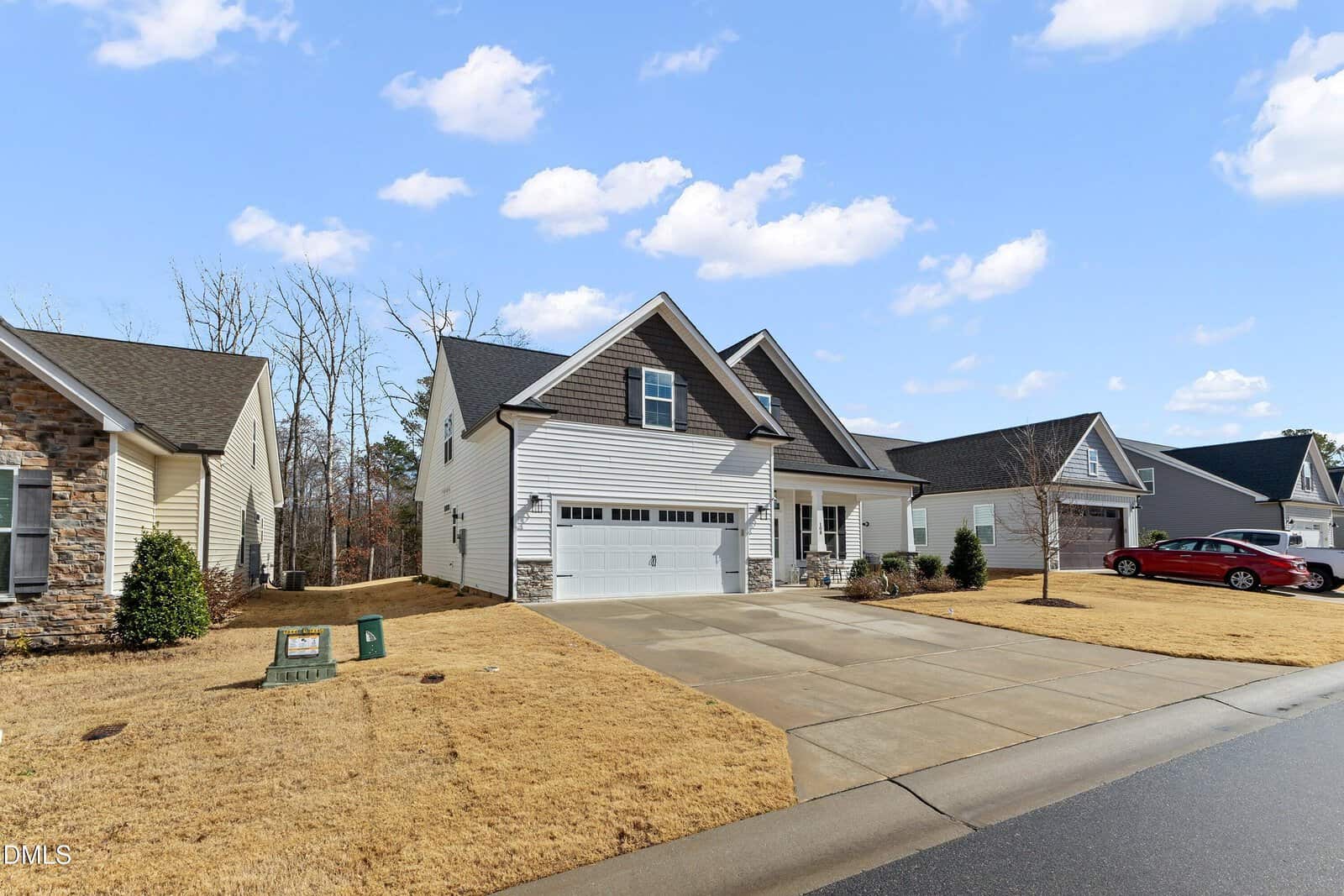 A modern suburban house with white siding, a gray roof, and an attached two-car garage sits on a dry, yellow lawn. Neighboring houses and a red car are visible. The sky is partly cloudy.