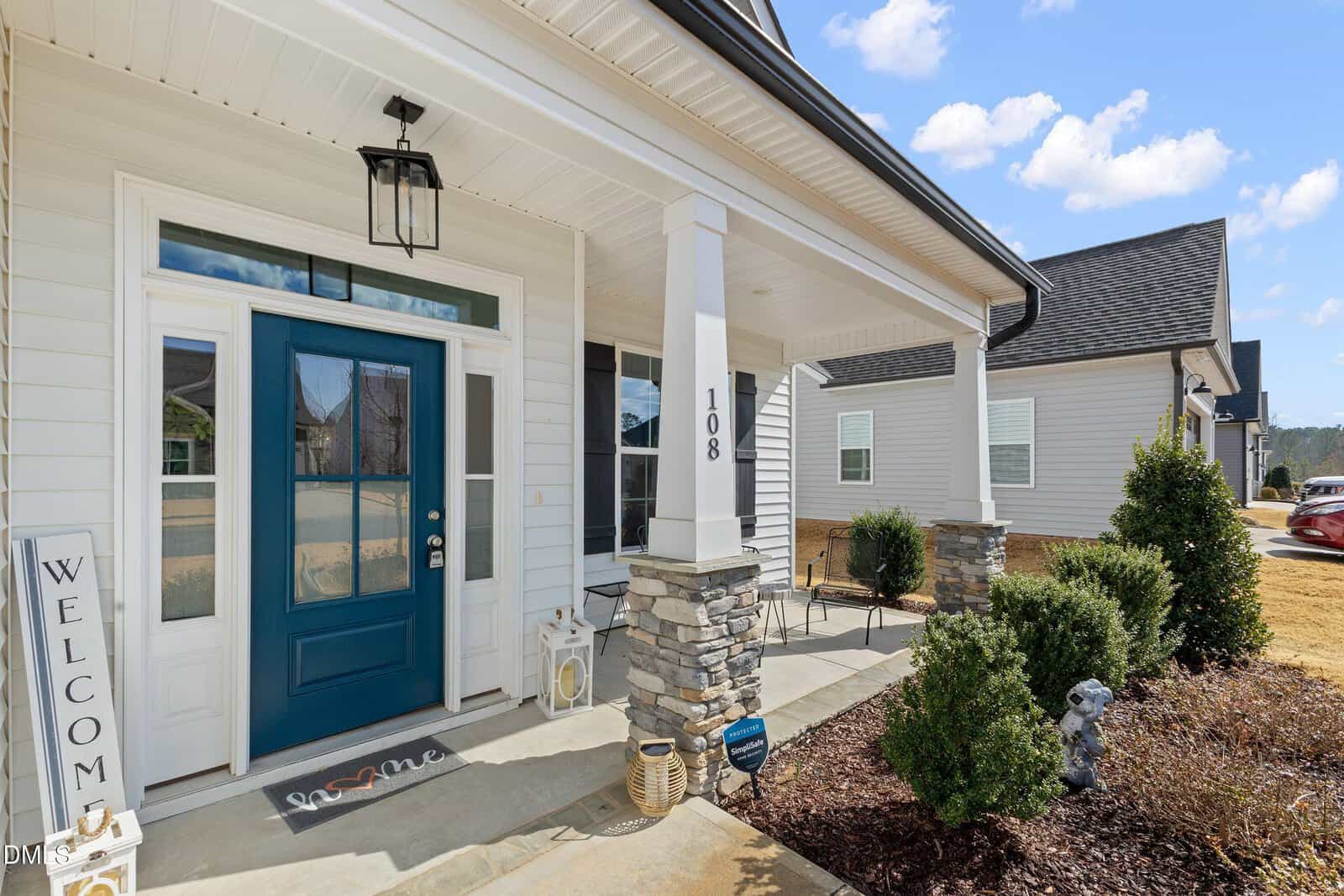 A house front porch with white siding, stone pillars, a blue front door with glass panels, a welcome sign, potted plants, and shrubbery. The house number 108 is visible on a column near the entrance.