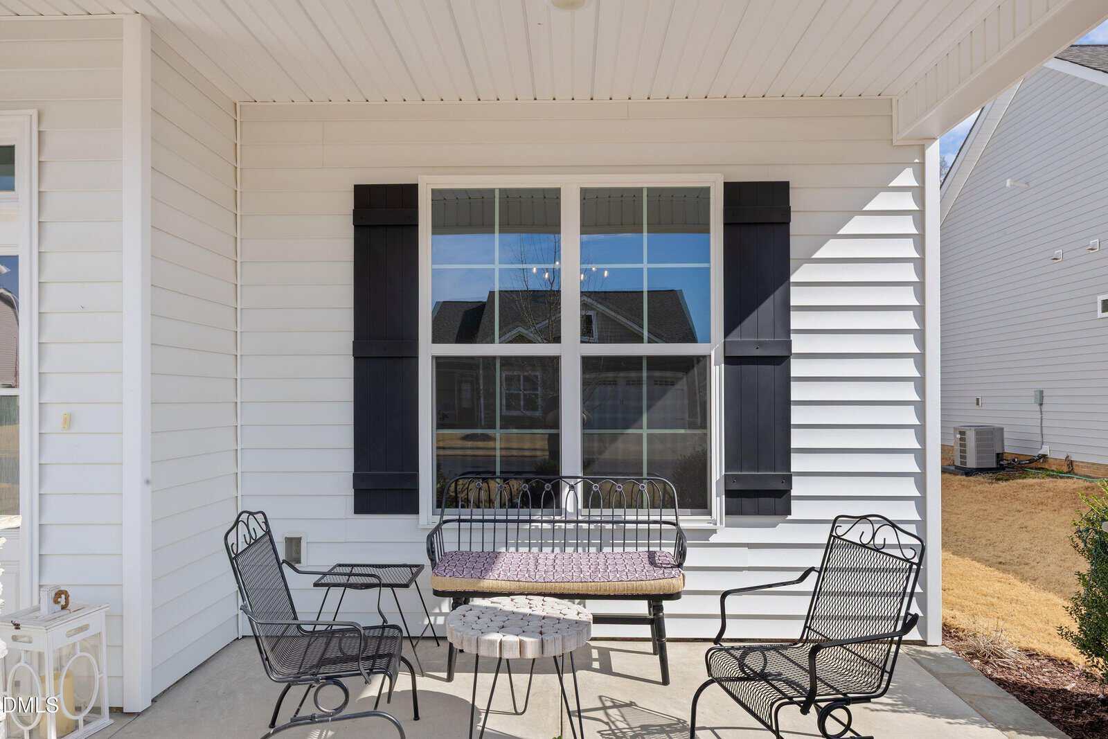 A patio with a small table, four black metal chairs, a black metal bench with a purple cushion, all set against a white house exterior with large windows and black shutters.