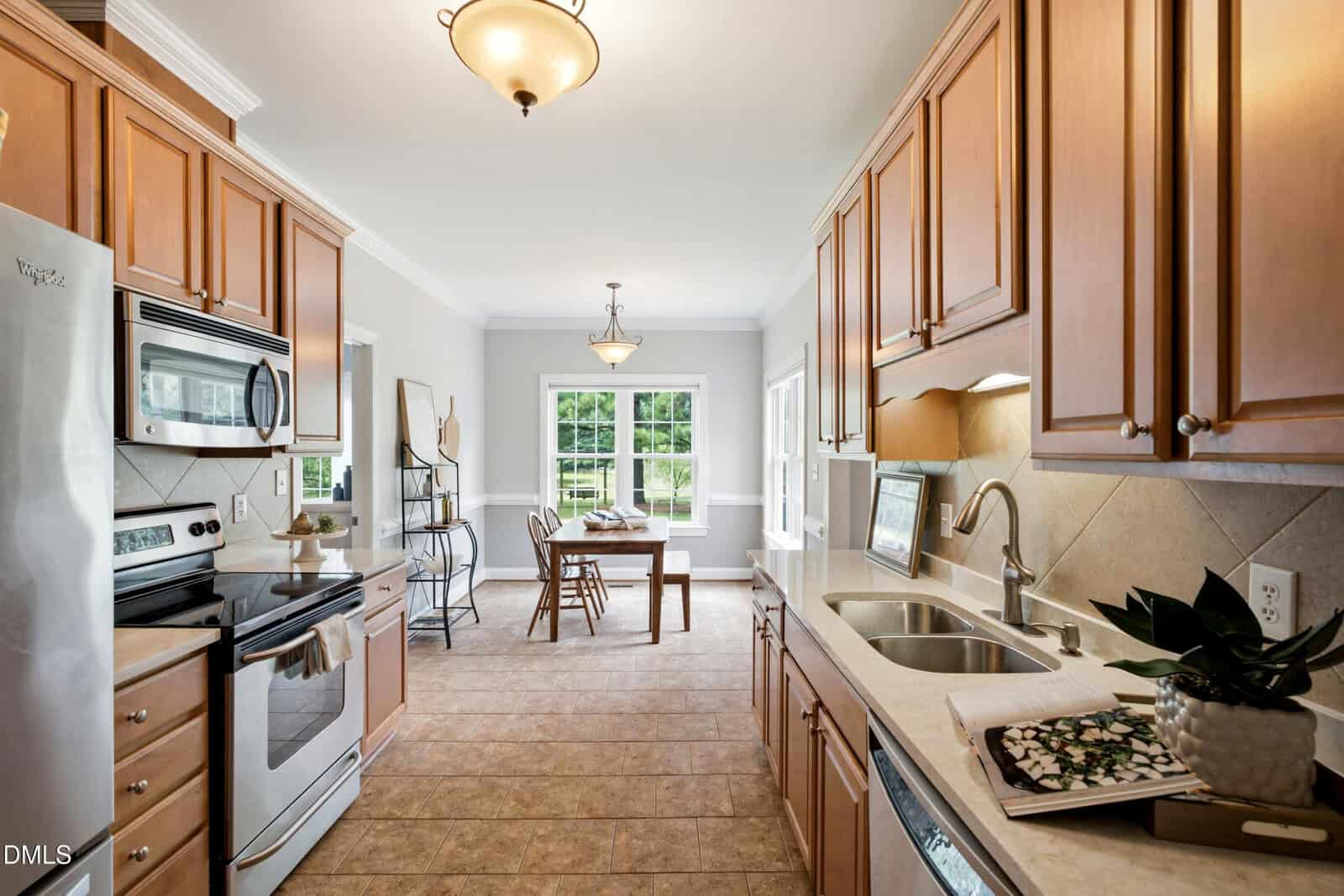 Bright kitchen with light wood cabinets, stainless steel appliances, double sink, and tiled floor. A dining table with two chairs sits in a sunny nook near large windows; a plant and open book are on the countertop.