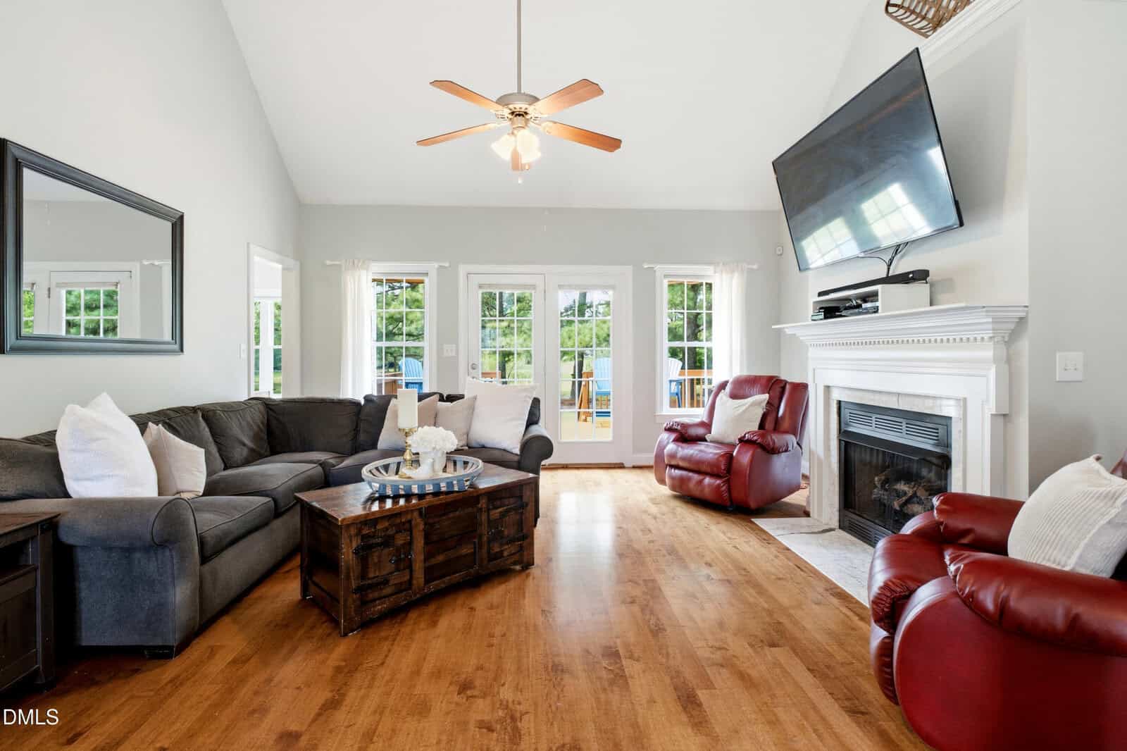Bright living room with a gray sectional sofa, two red recliners, a wooden coffee table, a fireplace with a mounted TV, large windows, and glass doors letting in natural light.