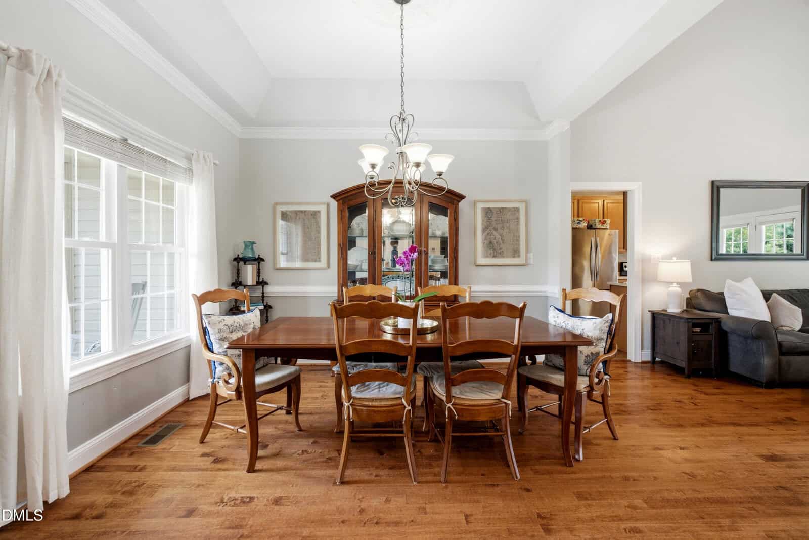 A dining room with a wooden table and six chairs, a chandelier overhead, and a glass-fronted hutch against the back wall. Hardwood floors, neutral walls, and windows let in natural light.