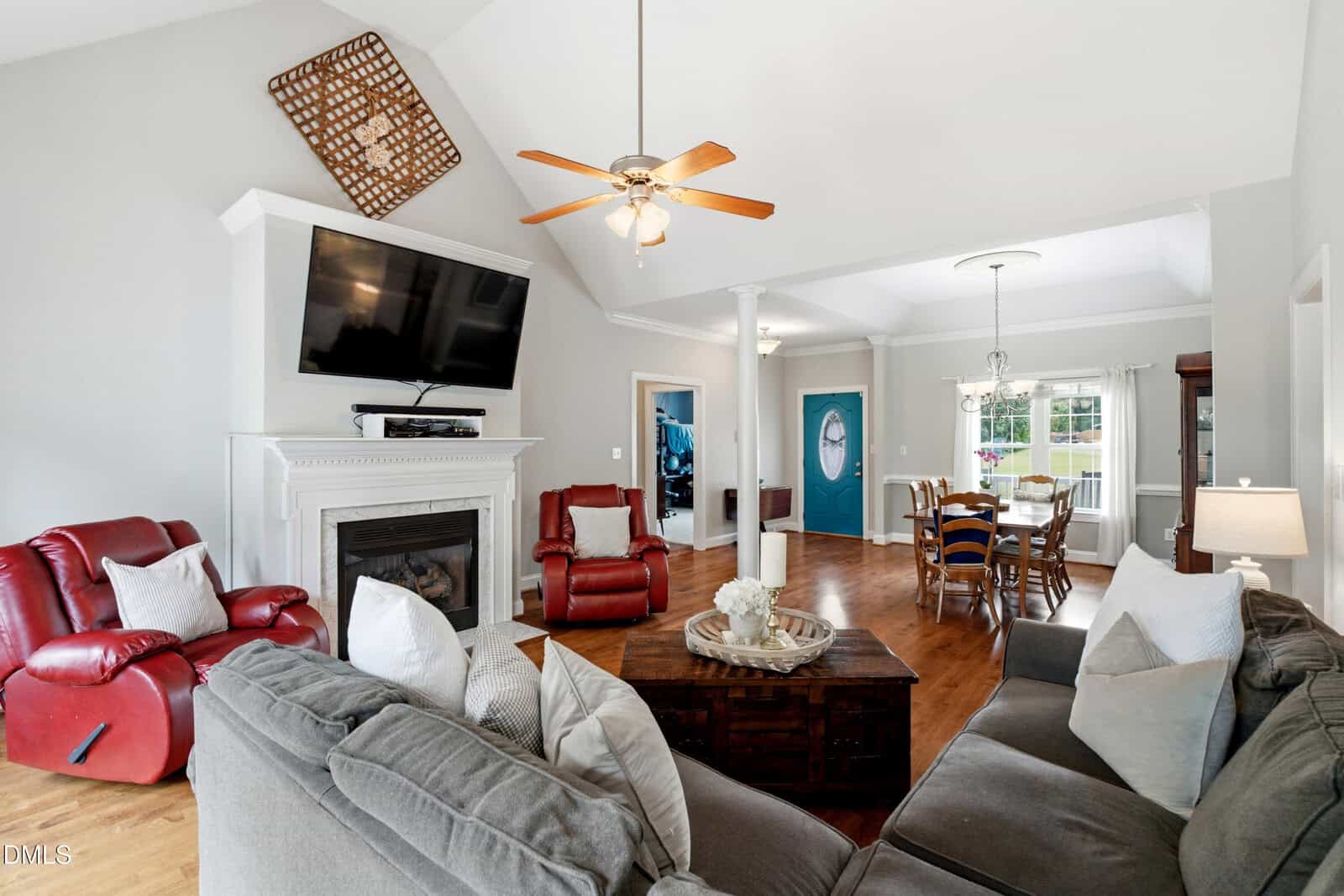 Bright living room with a gray sectional, red recliners, a fireplace with a TV above, wooden floors, and an open dining area with a blue accent wall and large windows letting in natural light.