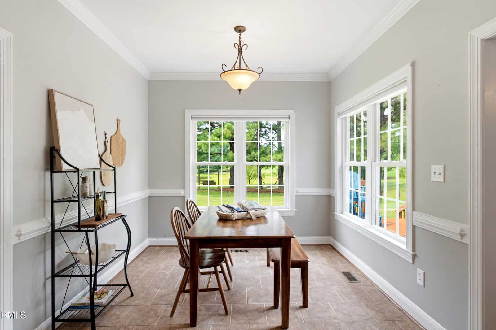 A dining area with a wooden table, two chairs, a bench, a metal shelf with decor, tile flooring, and large windows showing a green yard outside. A pendant light hangs from the ceiling. Walls are painted light gray.