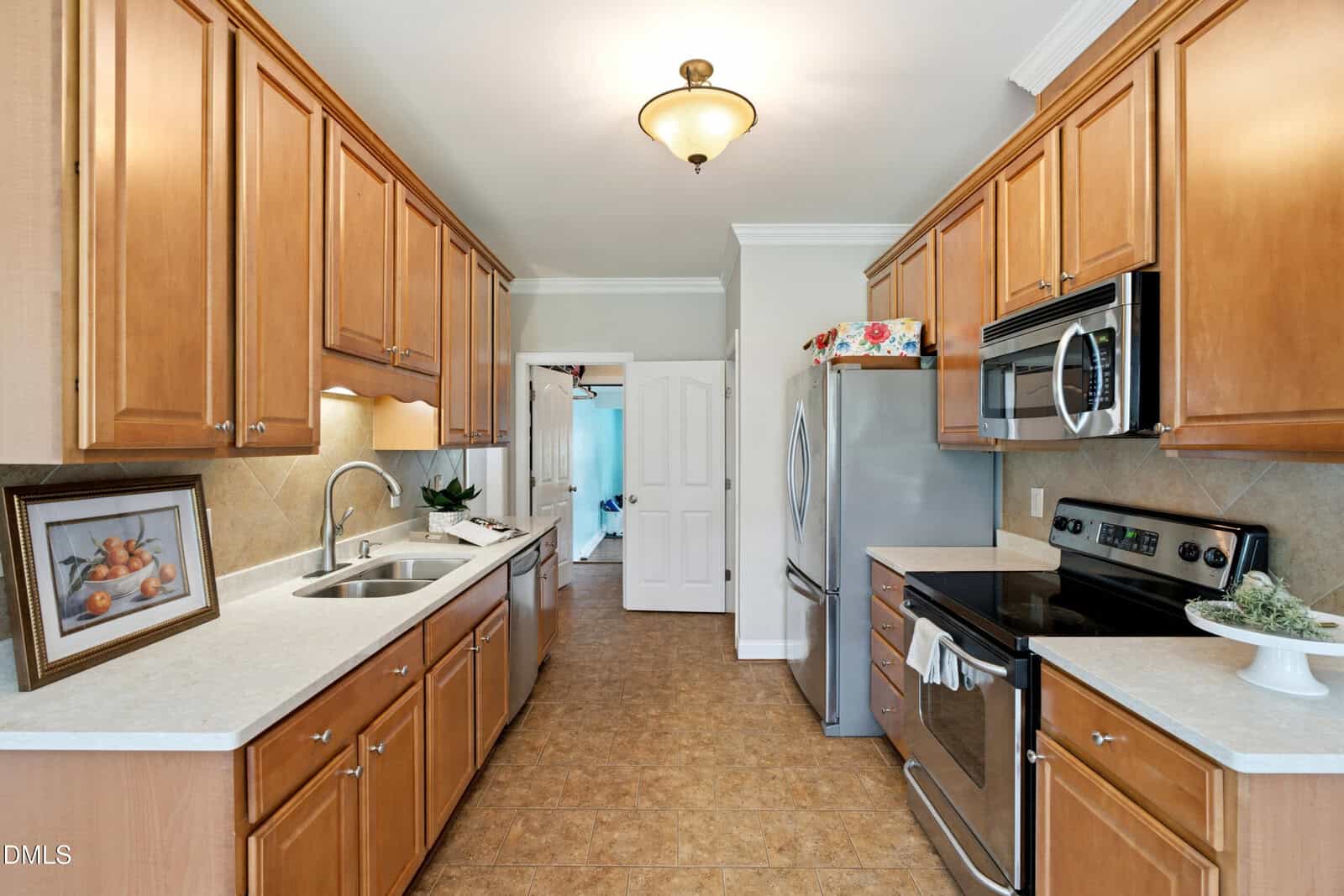 A galley kitchen with wood cabinets, stainless steel appliances, a double sink, light countertops, tile flooring, and neutral walls. A framed fruit picture leans against the backsplash on the left counter.