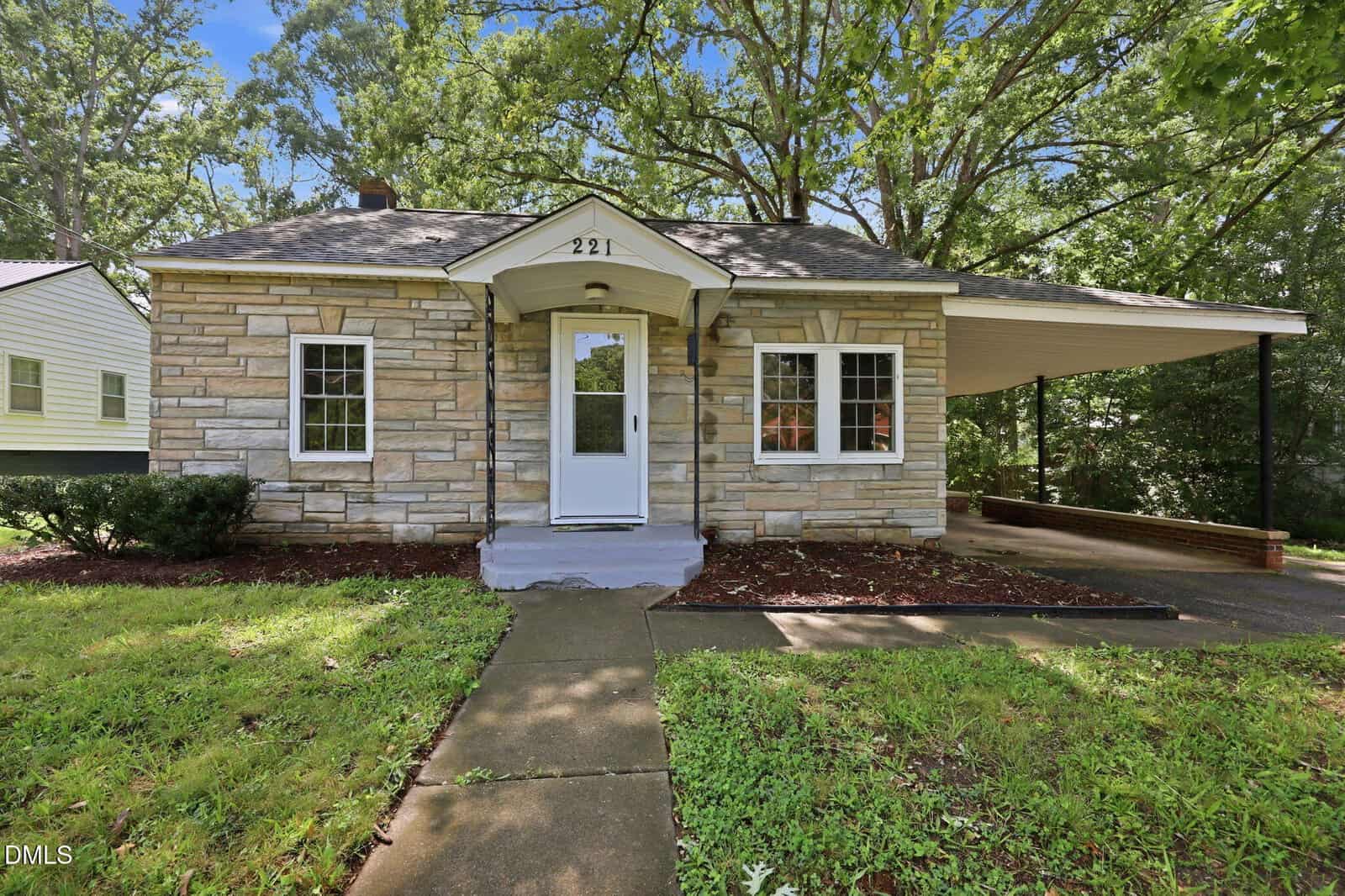 Charming single-story house with a stone exterior, small covered front porch, white door, two front windows, and an attached carport on the right. Located at 221 Zollicoffer Avenue in Henderson, the property features a yard with green grass and mature trees in the background.