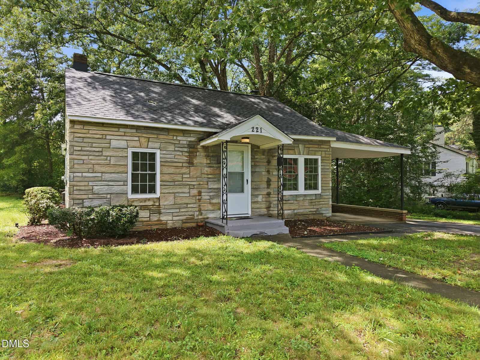 Small stone house with a covered front porch, white door, and two windows. Surrounded by grass and trees, with a paved walkway leading to the entrance. A driveway runs along the right side of the house.