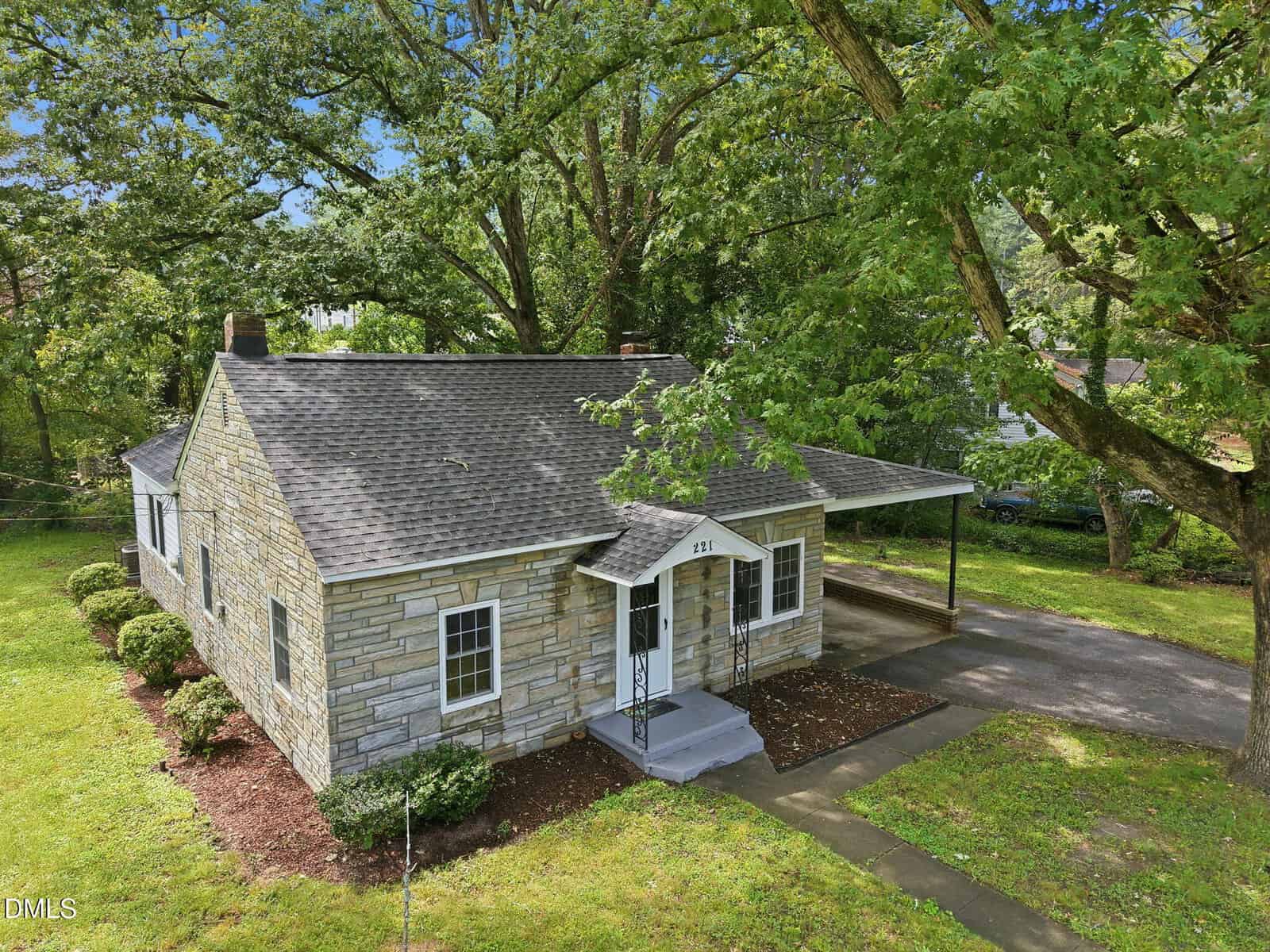 Single-story stone house with a gray shingled roof, covered carport, small front porch, and surrounded by green grass and large trees. A concrete walkway leads to the front door.