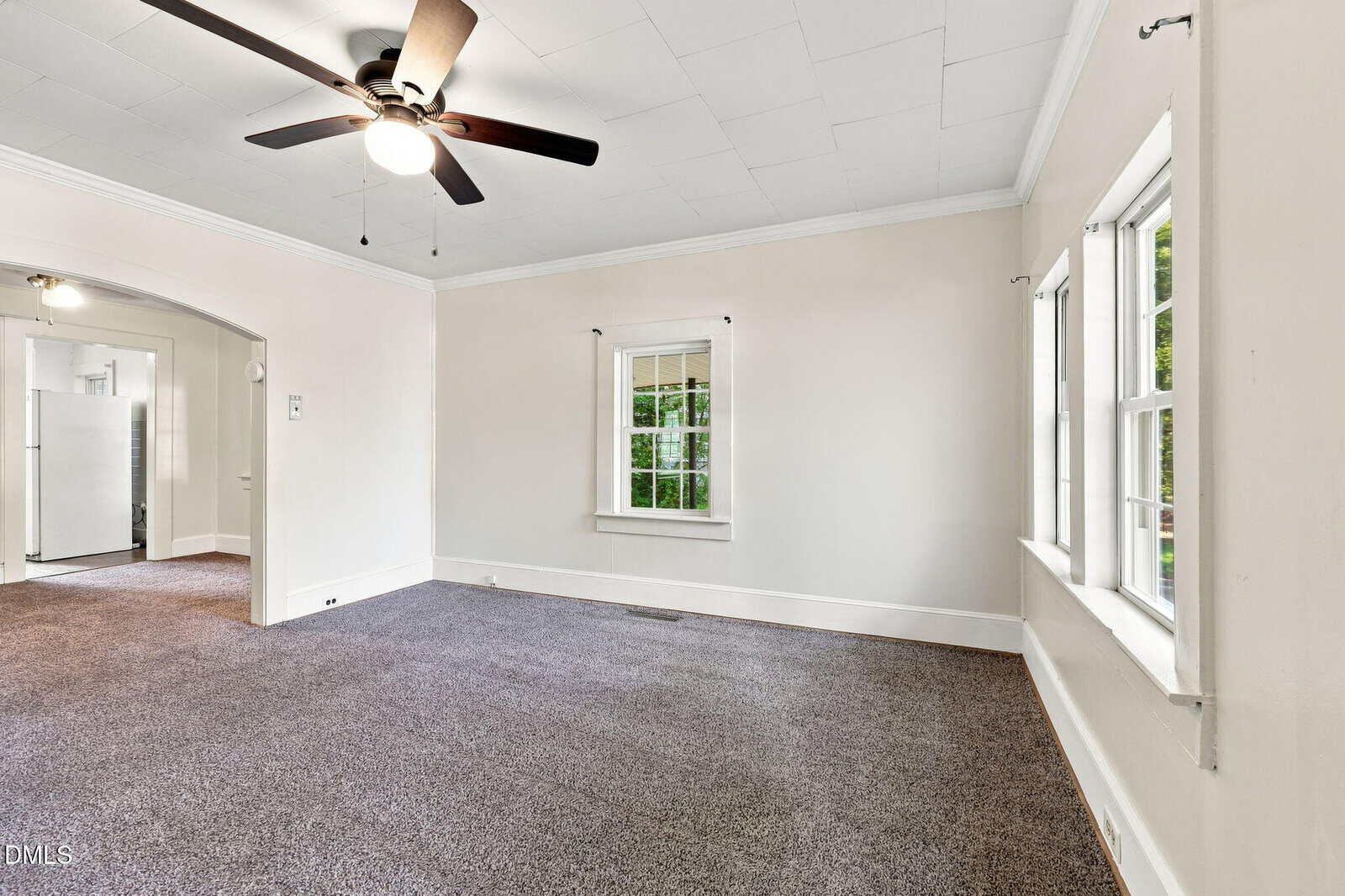 Unfurnished living room with gray carpet, white walls, a ceiling fan with lights, and three windows letting in natural light. An arched doorway leads to a kitchen area with visible refrigerator.