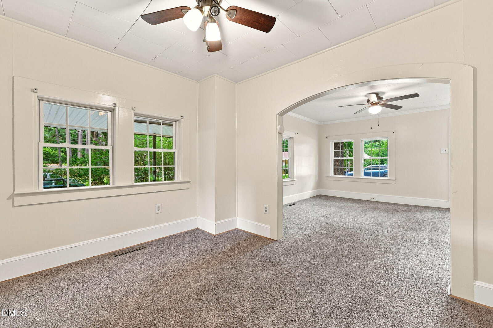 A carpeted living and dining area with beige walls, two ceiling fans, multiple windows with views of greenery, and an arched opening dividing the rooms.