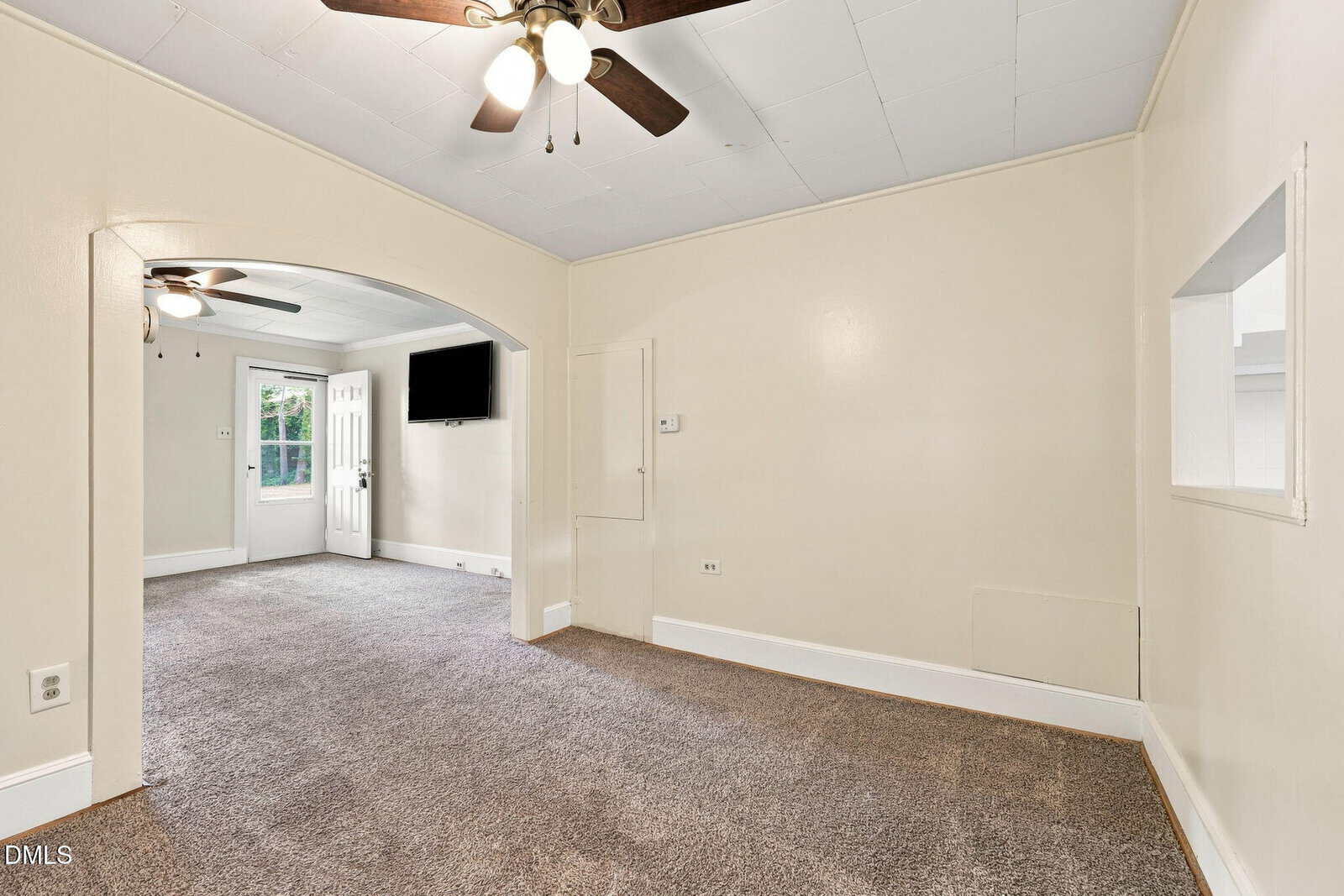 A bright room with beige walls, carpeted floor, ceiling fans, a wall-mounted TV, and an open doorway leading to another area with a window and white door.