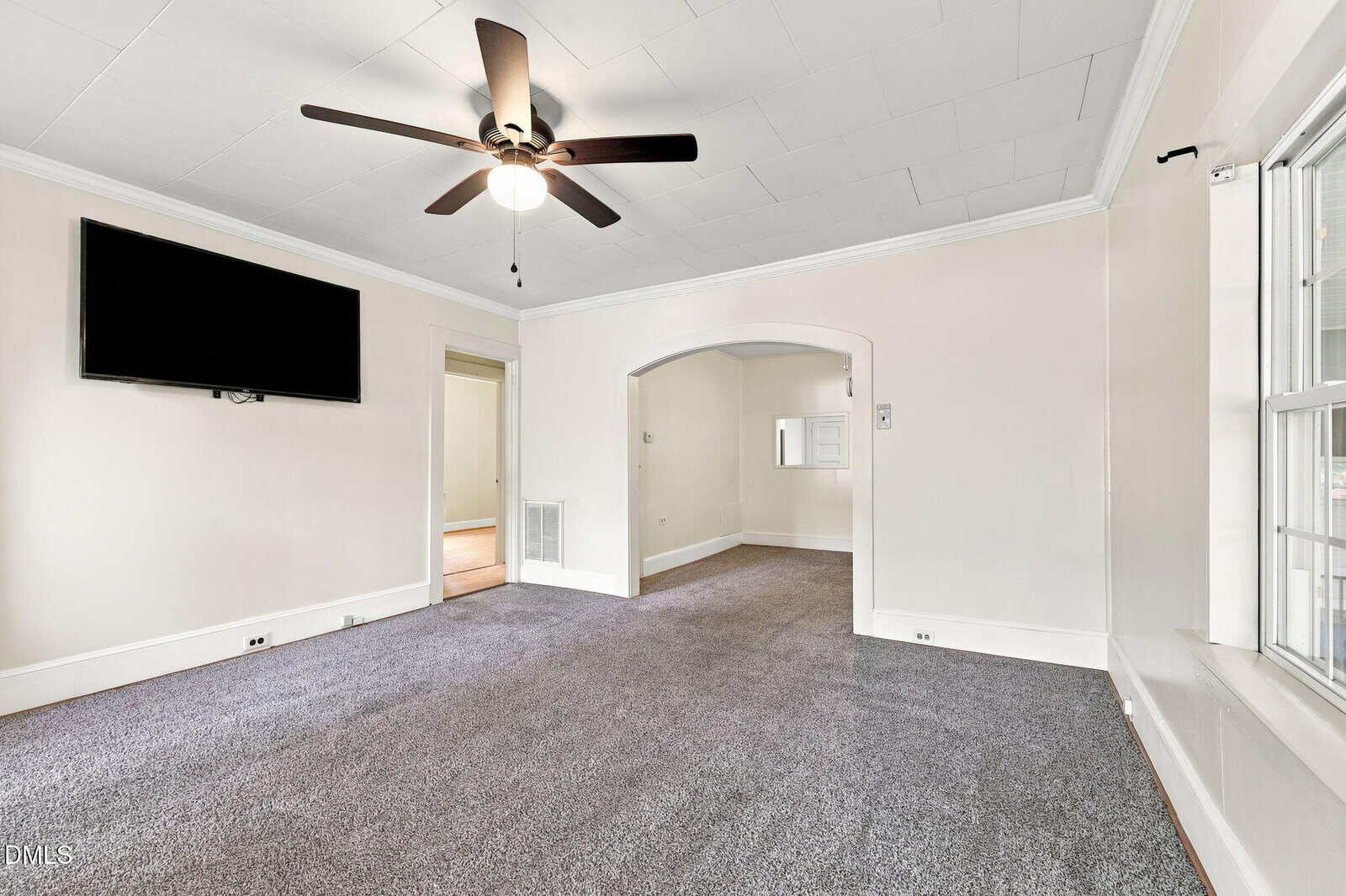 Spacious living room with gray carpet, ceiling fan, large wall-mounted TV, and white walls. An arched doorway leads to another room, and there is a large window on the right.