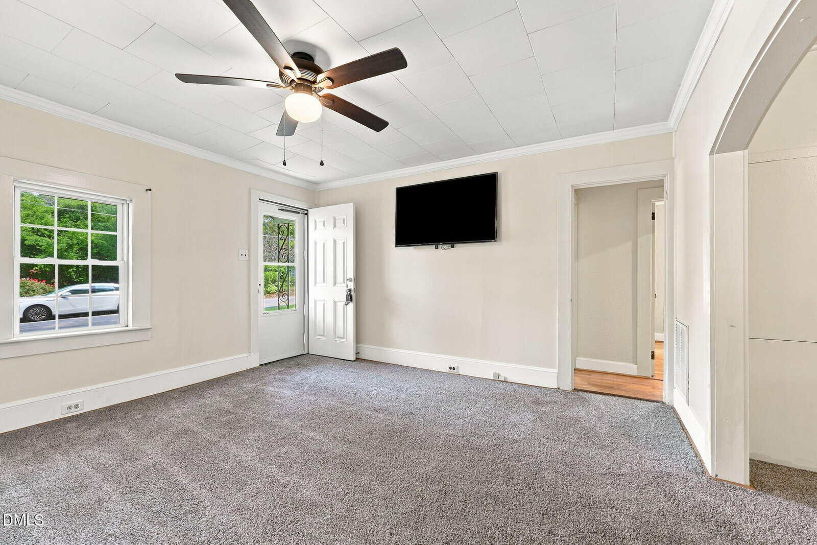 Bright living room with a ceiling fan, gray carpet, wall-mounted TV, and white walls. There’s a window and door to the outside, plus an open doorway to a hallway and an arched entry to another room.