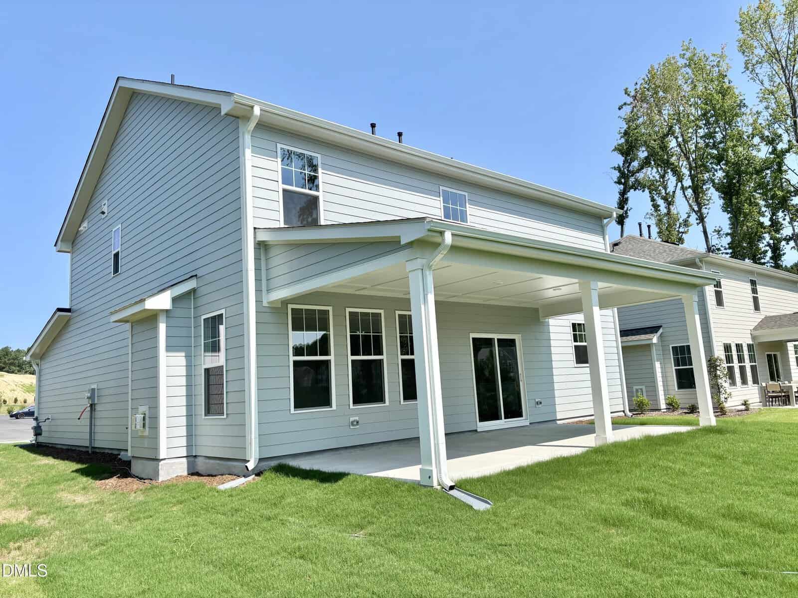 Two-story house with light gray siding and white trim, featuring multiple windows and a large covered back patio. The yard is grassy and well-maintained, with neighboring houses visible in the background.