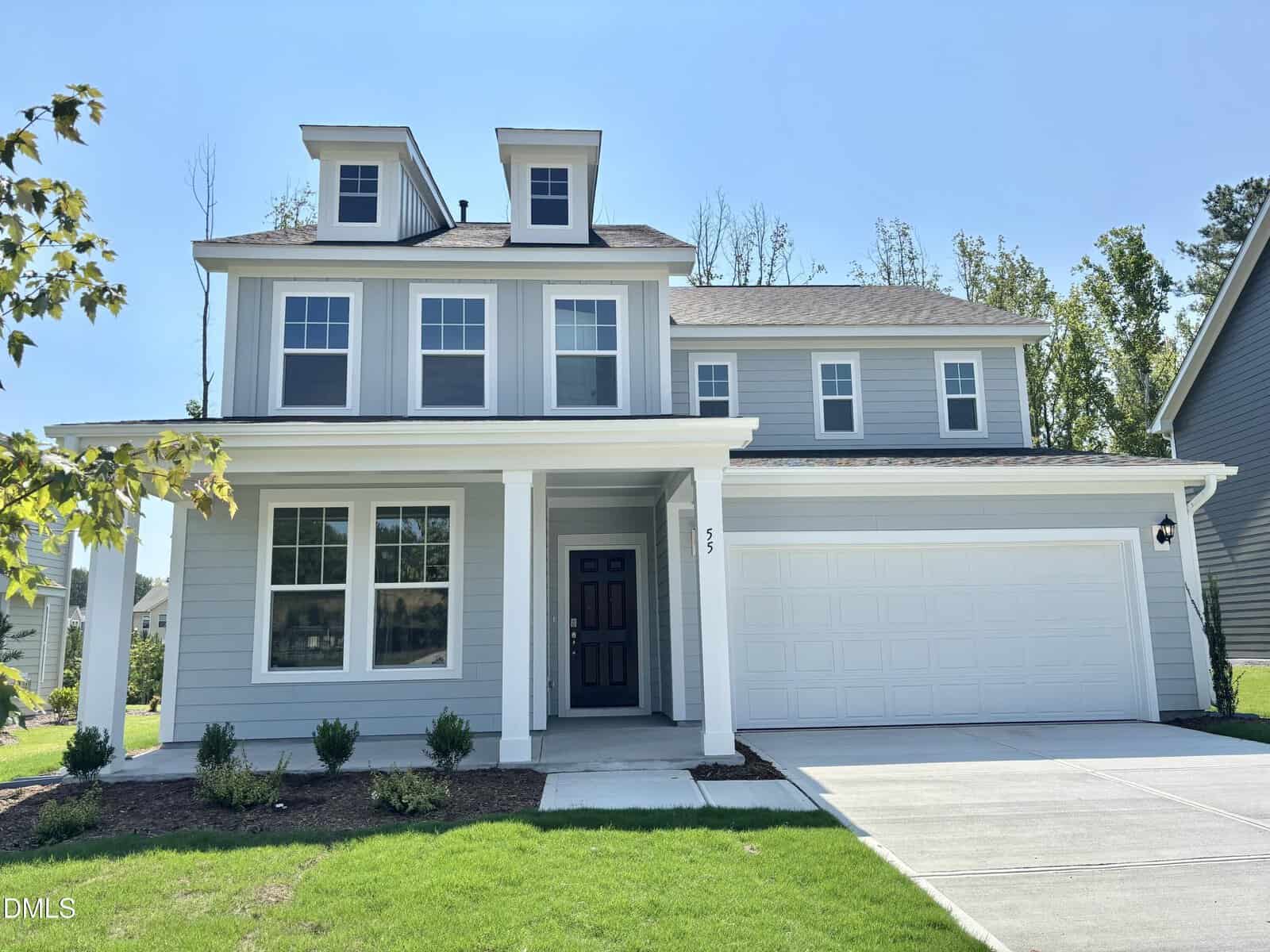 Two-story modern gray house with white trim, covered front porch, double garage door, and a manicured lawn. The house has multiple windows and dormers on the roof. There are small shrubs planted along the front.