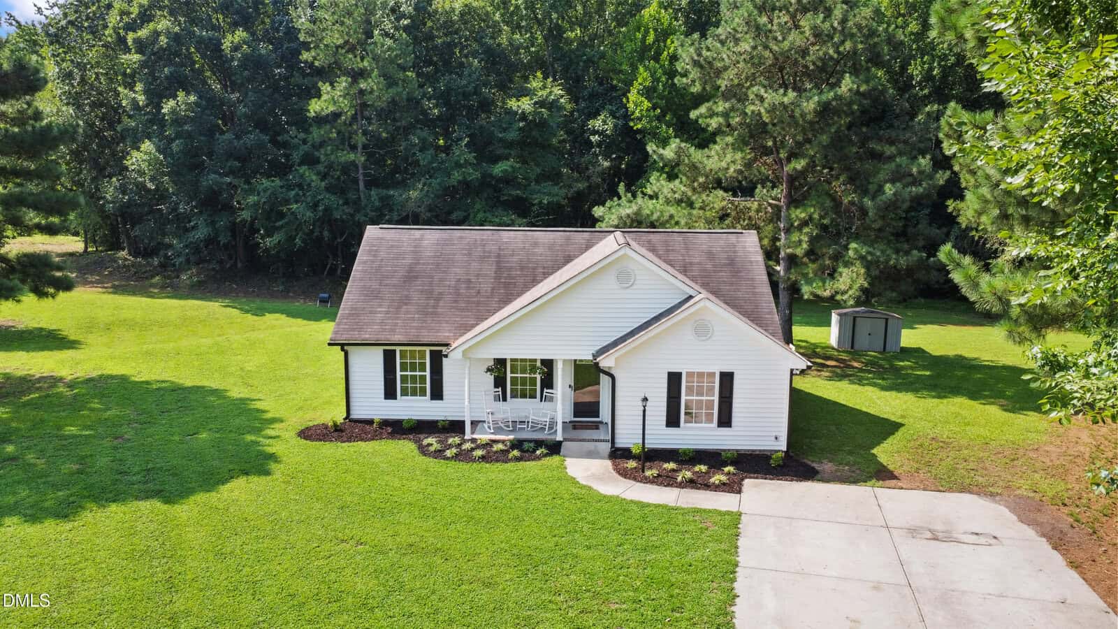 A small, single-story white house with a covered front porch, dark roof, two front windows, and a driveway, surrounded by green grass, trees, and a backyard storage shed.