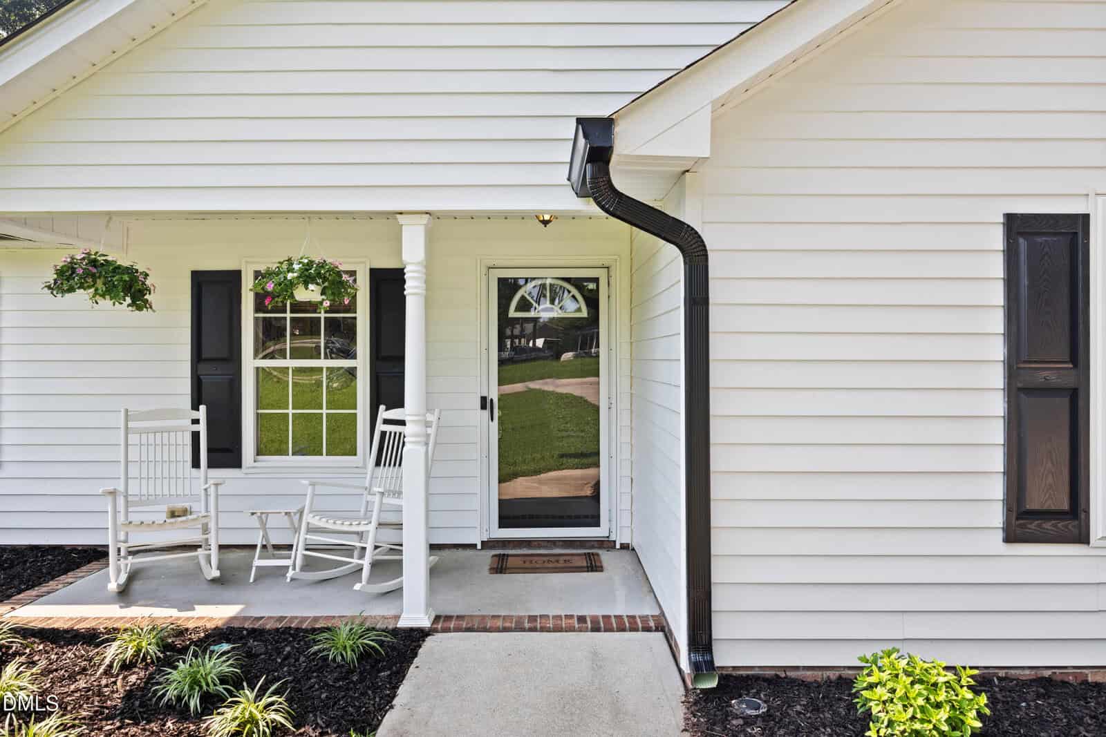 White house exterior with black shutters and a covered front porch. Two white rocking chairs and hanging plants are on the porch. A glass front door with a welcome mat leads to a walkway. Neatly landscaped garden beds are visible.