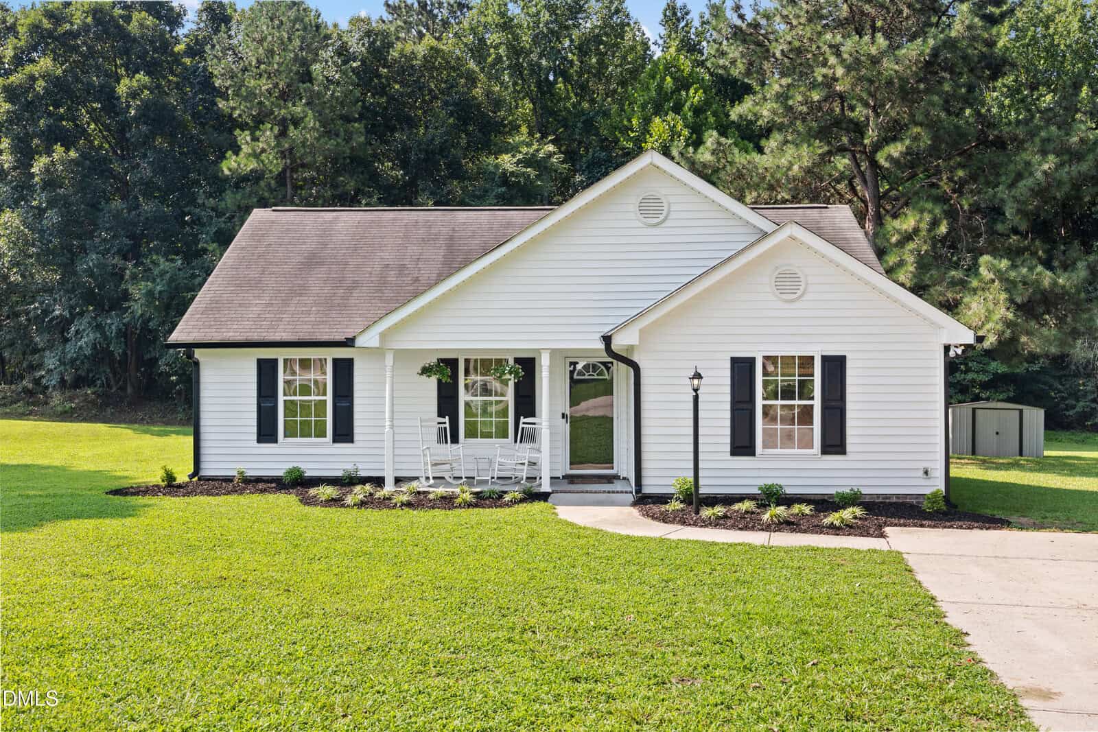 A single-story white house with black shutters, a front porch with two rocking chairs, a manicured lawn, and a curved driveway awaits you at 65 Rainwood Court in Louisburg, surrounded by picturesque trees in the background.