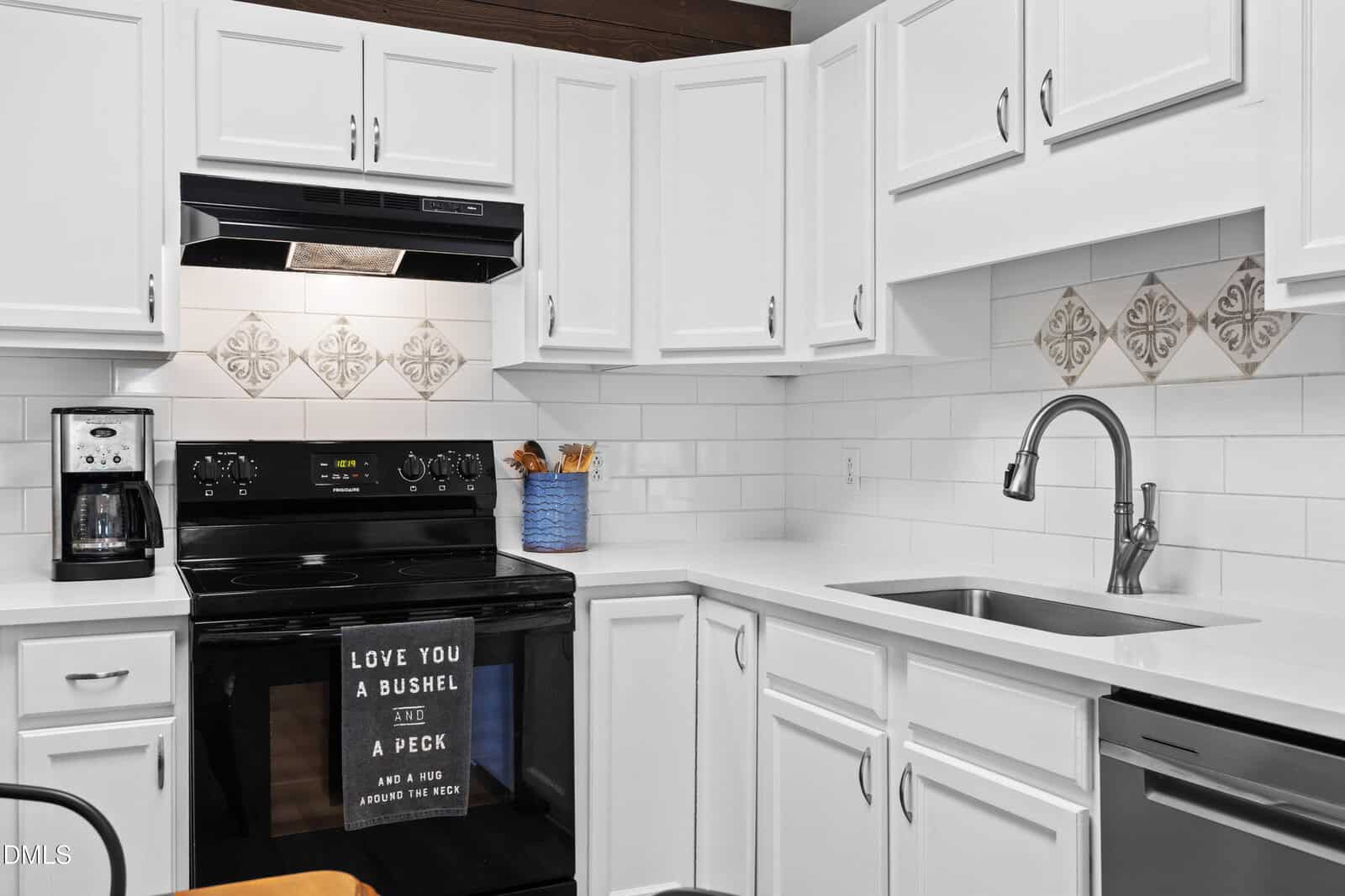 A modern white kitchen with black appliances, a coffee maker, a sink, and a stove. A kitchen towel with text hangs on the oven. Decorative tiles are above the stove. A utensil holder sits on the counter.