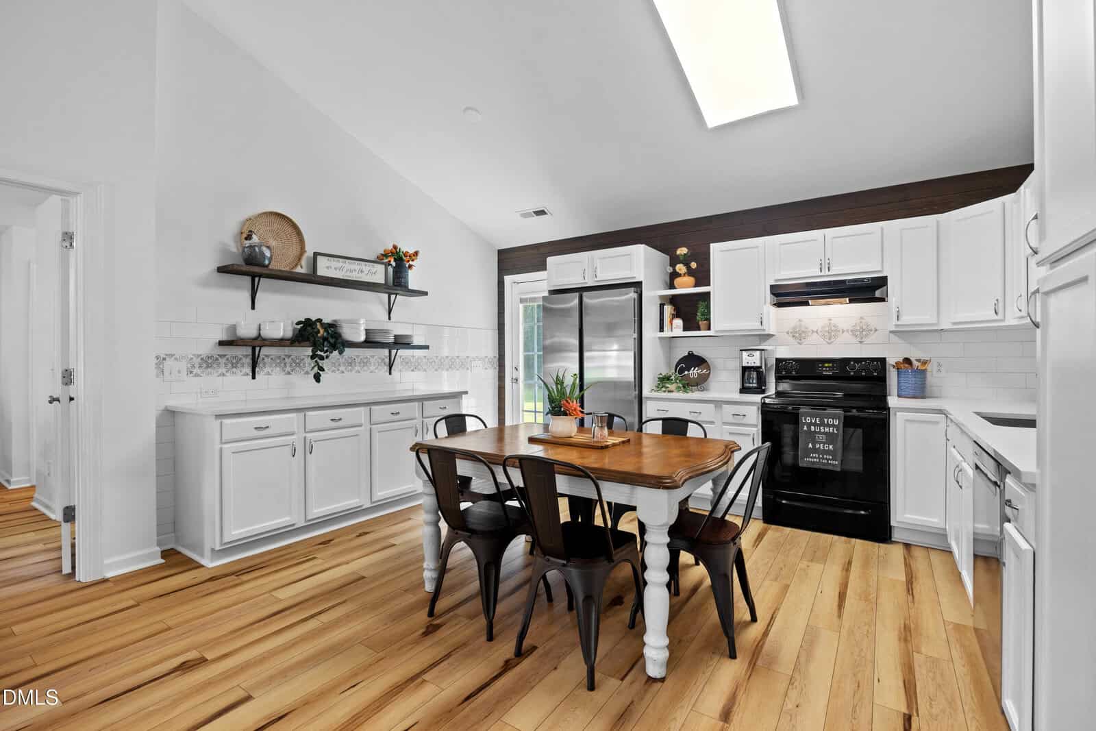 Bright kitchen in Louisburg with white cabinets, wood floors, black appliances, a wooden dining table with six metal chairs, floating shelves with decor, and a large fridge. Skylight and French doors provide natural light in this inviting home at 65 Rainwood Court.