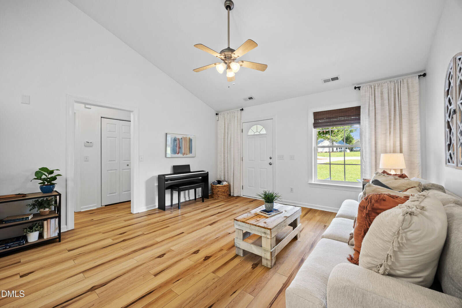 A bright living room with light wood floors, a beige sofa with pillows, a rustic coffee table, a piano, a ceiling fan, white walls, and a window with white curtains showing green grass and trees outside.