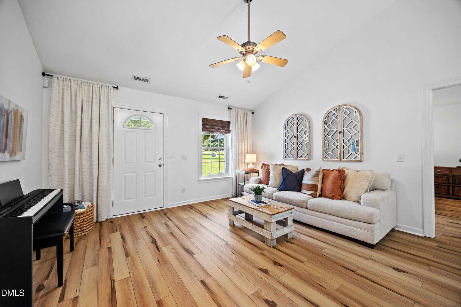 Bright living room with wood floors, a white sofa with colorful pillows, a coffee table, two decorative mirrors on the wall, a piano, and a ceiling fan. Natural light enters through a window and glass door.