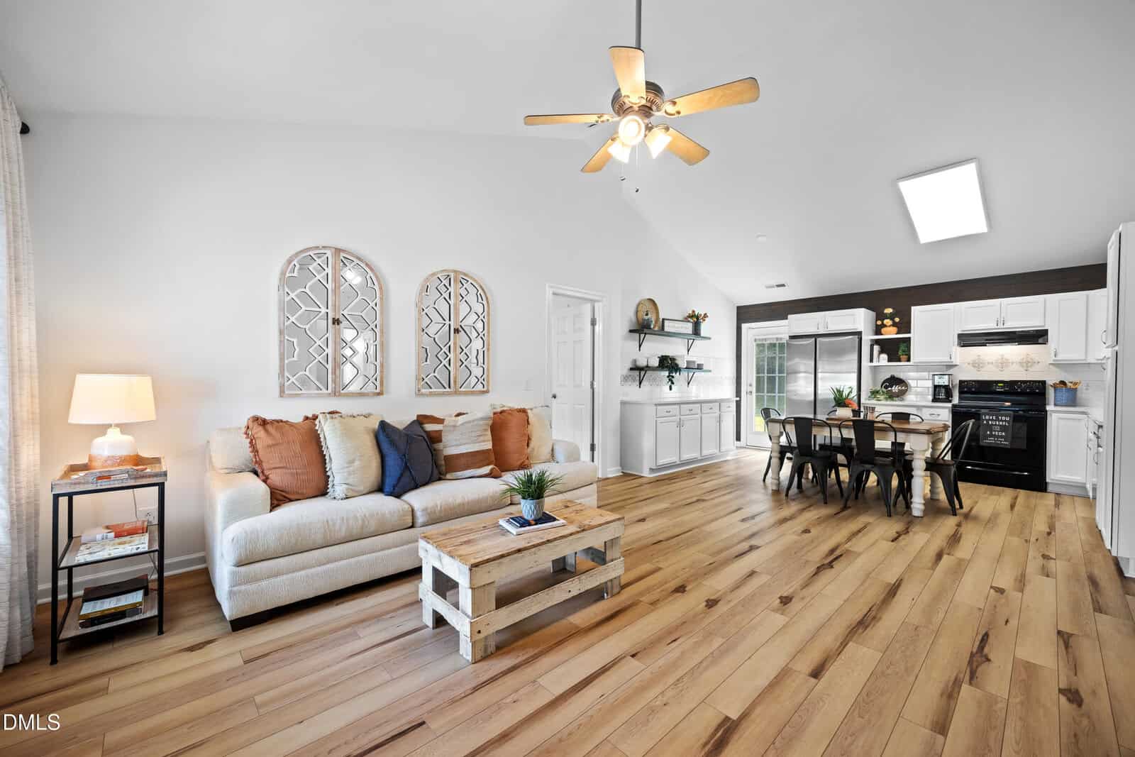 Bright open-concept living room and kitchen with light wood floors, white walls, a beige sofa, wooden coffee table, dining table, ceiling fan, and modern decor. Natural and artificial lighting fill the space.