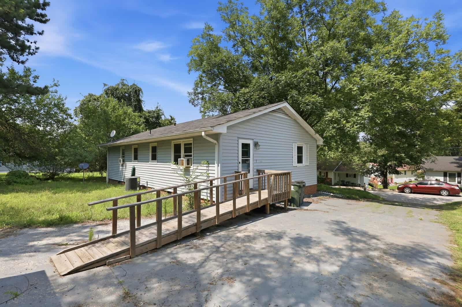 Single-story house with light-colored siding, a wooden wheelchair ramp leading to the front door, surrounded by trees and grass. A red car is parked on the street in the background on a sunny day.