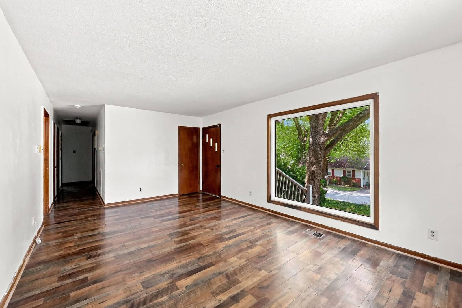 Empty room with white walls, brown wooden doors, and dark wood floors. A large window on the right overlooks a tree and street. Light from the window brightens the room. A hallway extends into the distance on the left.