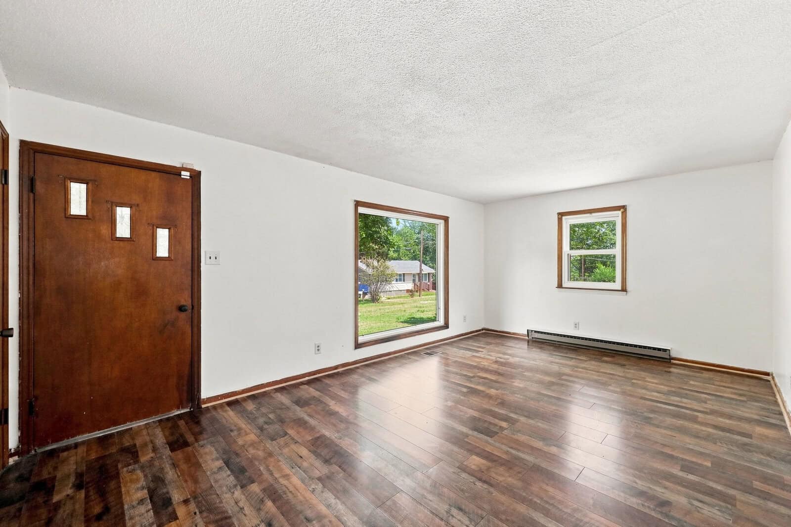 Empty living room with white walls, dark wood floor, a brown front door, a large picture window, and a smaller window on the adjacent wall. Baseboard heater runs along one wall. Trees and houses are visible outside.