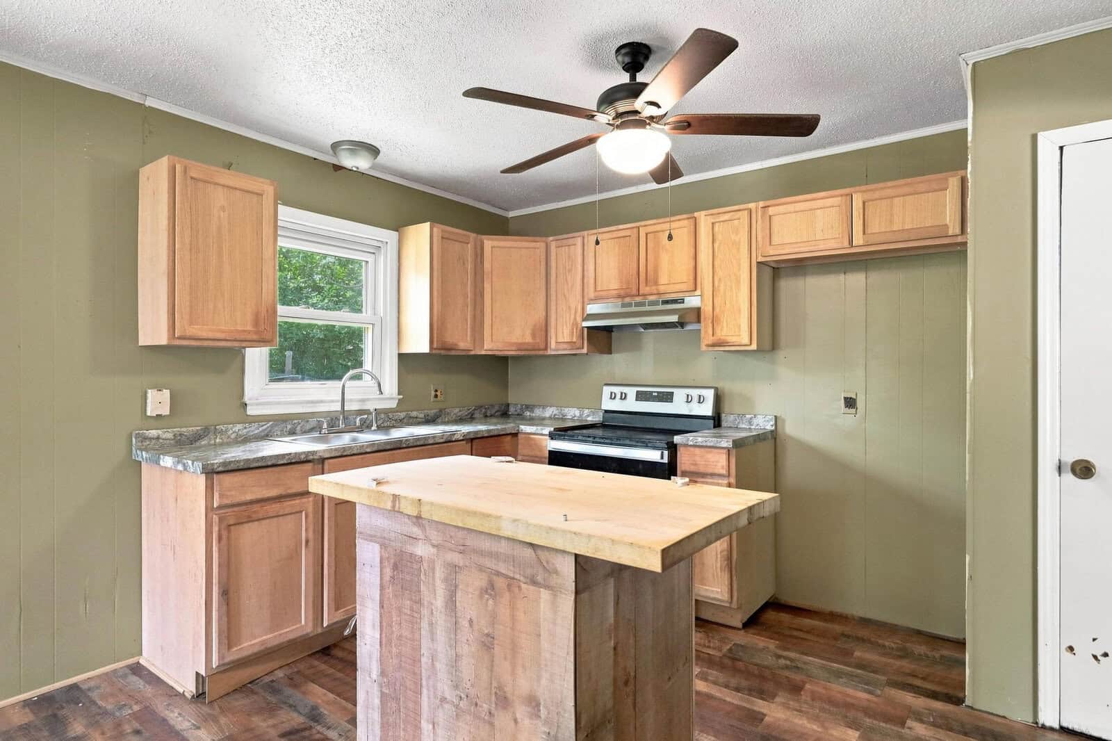 A small kitchen with light wood cabinets, a marble-patterned countertop, a stainless steel electric stove, a ceiling fan with a light, and a freestanding wooden island on dark wood flooring.