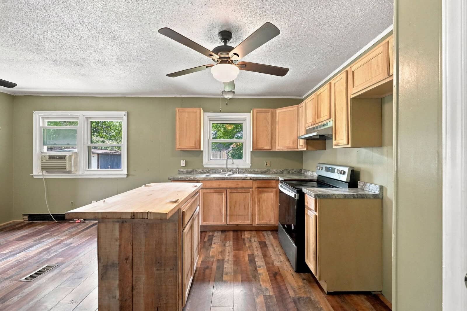 Kitchen with wood cabinets, stainless steel appliances, a central island, wood flooring, ceiling fan, and two windows; one has a window air conditioner.