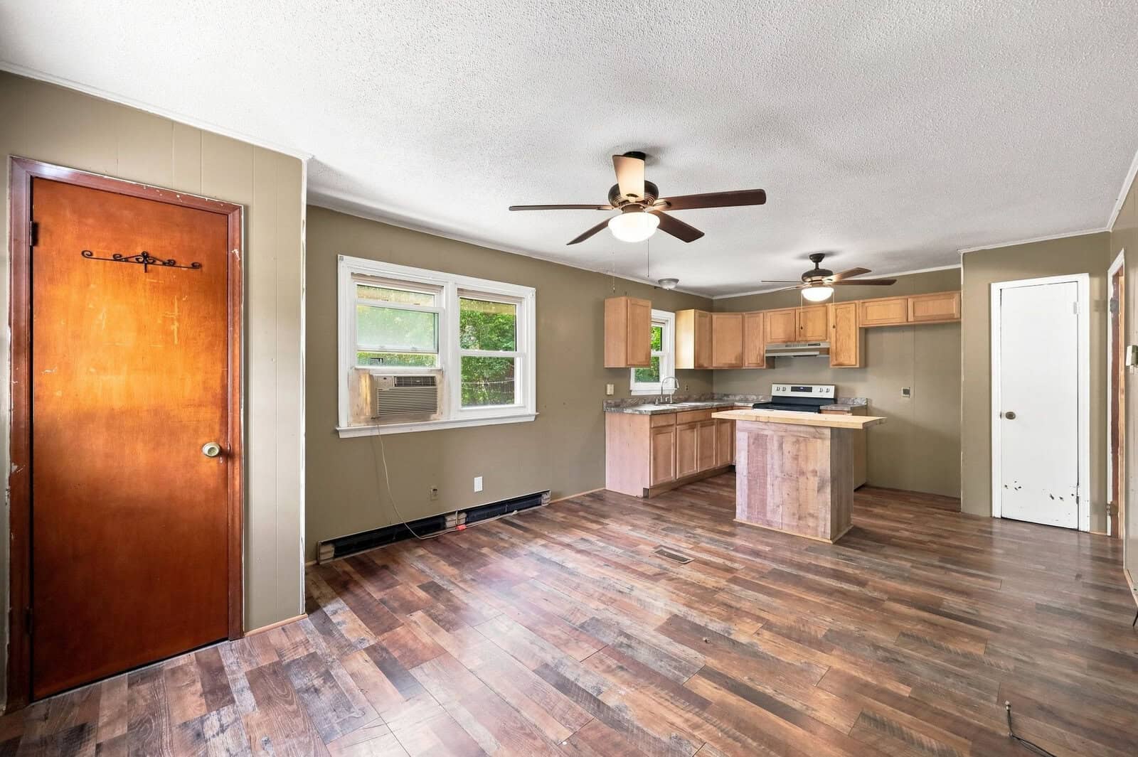 Open kitchen and living area with wood floors, brown cabinets, two ceiling fans, a window with AC unit, and a brown wooden door. The space has neutral walls and natural light coming through the windows.