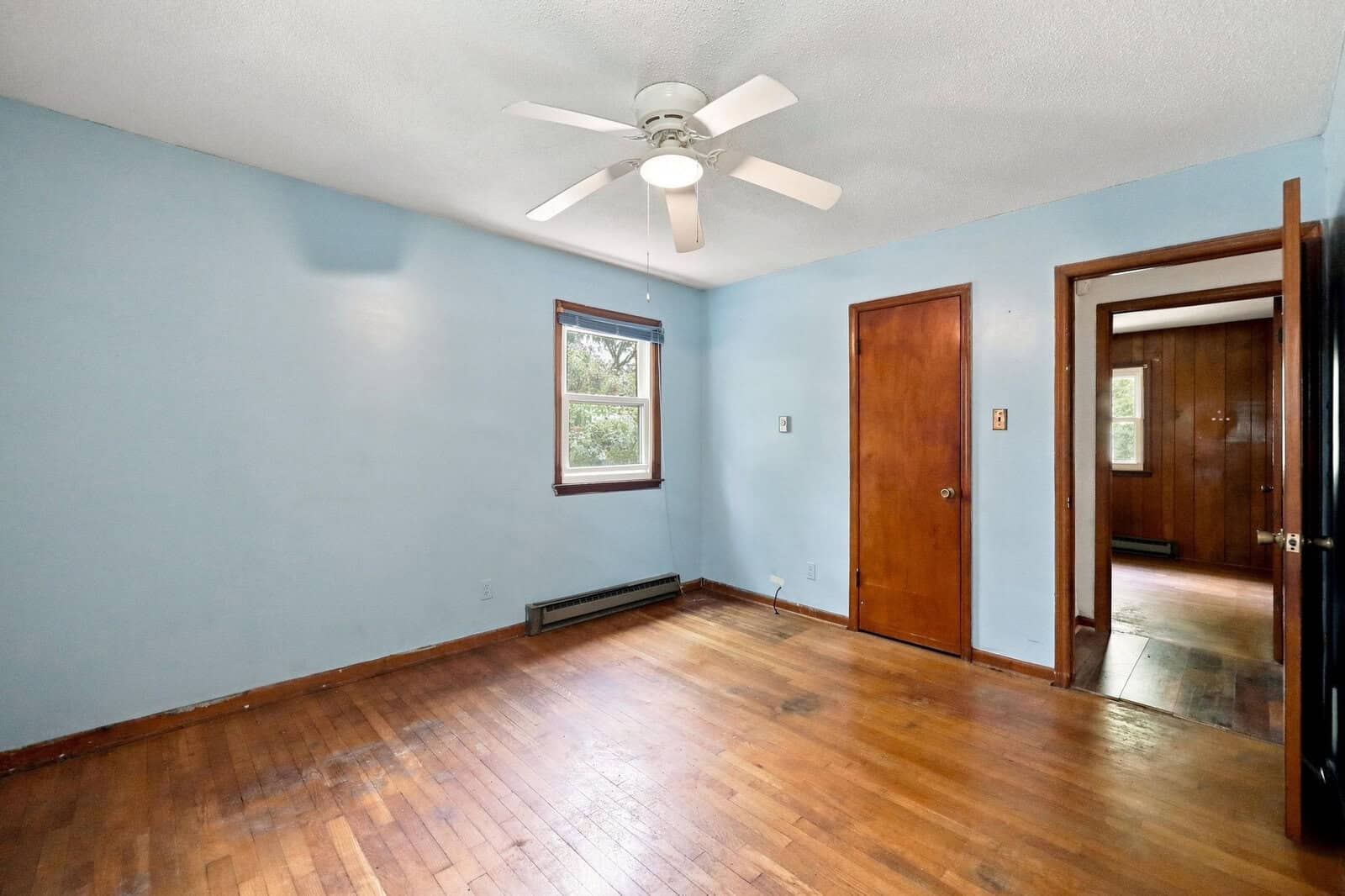 Empty room with light blue walls, hardwood floors, a white ceiling fan, two wooden doors, a window with natural light, and a baseboard heater along one wall.