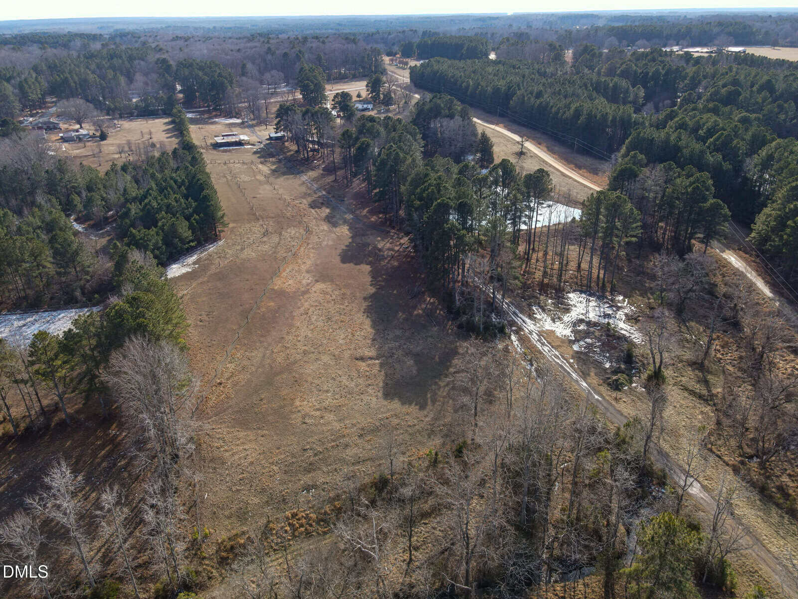 Aerial view of a rural landscape featuring open fields, clusters of trees, a few houses, dirt roads, and a small pond, with forests in the background. The scene appears to be in late winter or early spring.