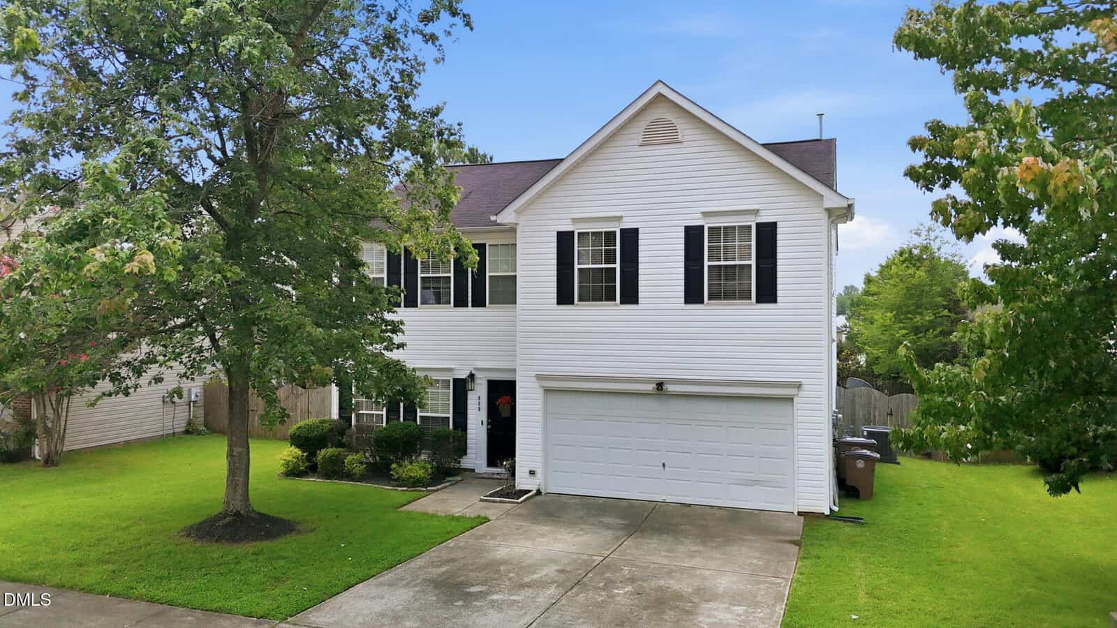 Two-story white house with black shutters, attached two-car garage, concrete driveway, and a well-kept front lawn. Trees and shrubs are present along the front and sides. The sky is clear with a few clouds.