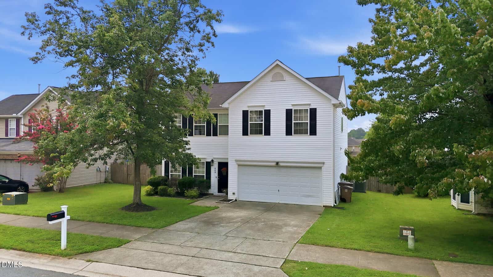 Two-story white house with black shutters and a double garage, set on a green lawn with a large tree in front. A white mailbox and concrete driveway lead to the house. Neighboring homes are visible on each side.