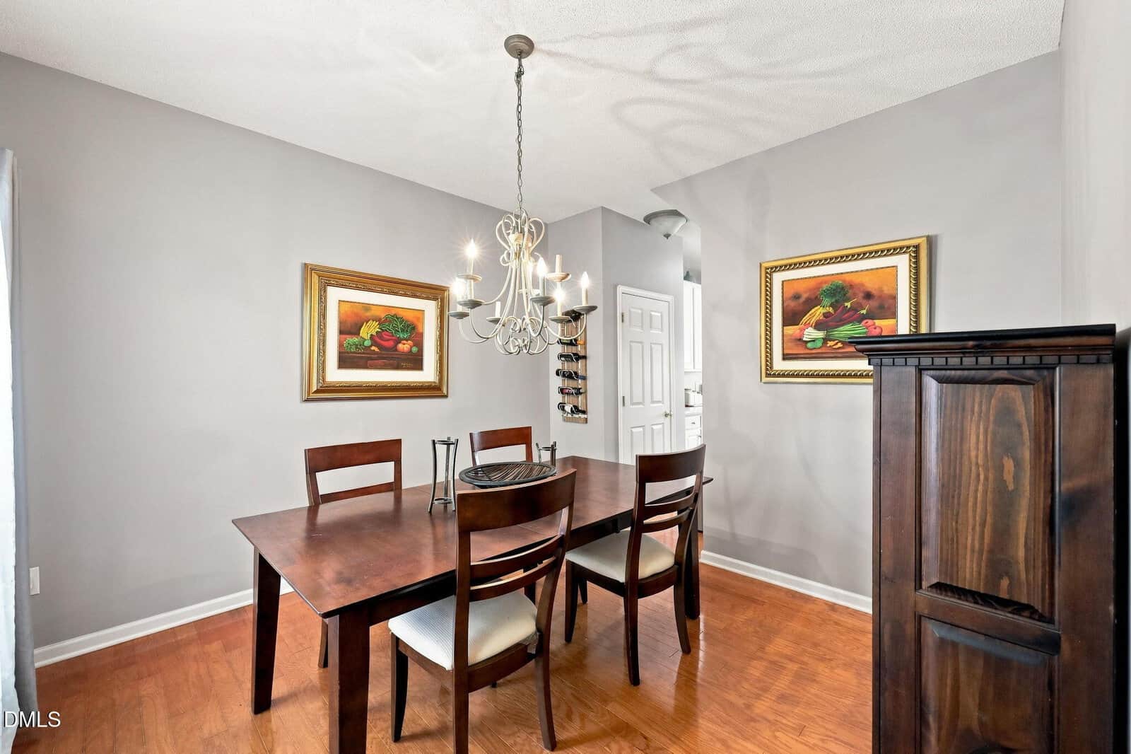 A dining room with a dark wood table and four chairs, a chandelier above, two framed paintings on the walls, a tall wooden cabinet, and a small wine rack near a white door. The walls are light gray and the floor is wood.