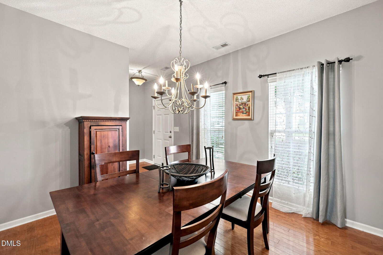 Dining room with a dark wooden table and chairs, modern chandelier, large windows with white curtains, gray walls, wooden cabinet, framed art, and hardwood floor.