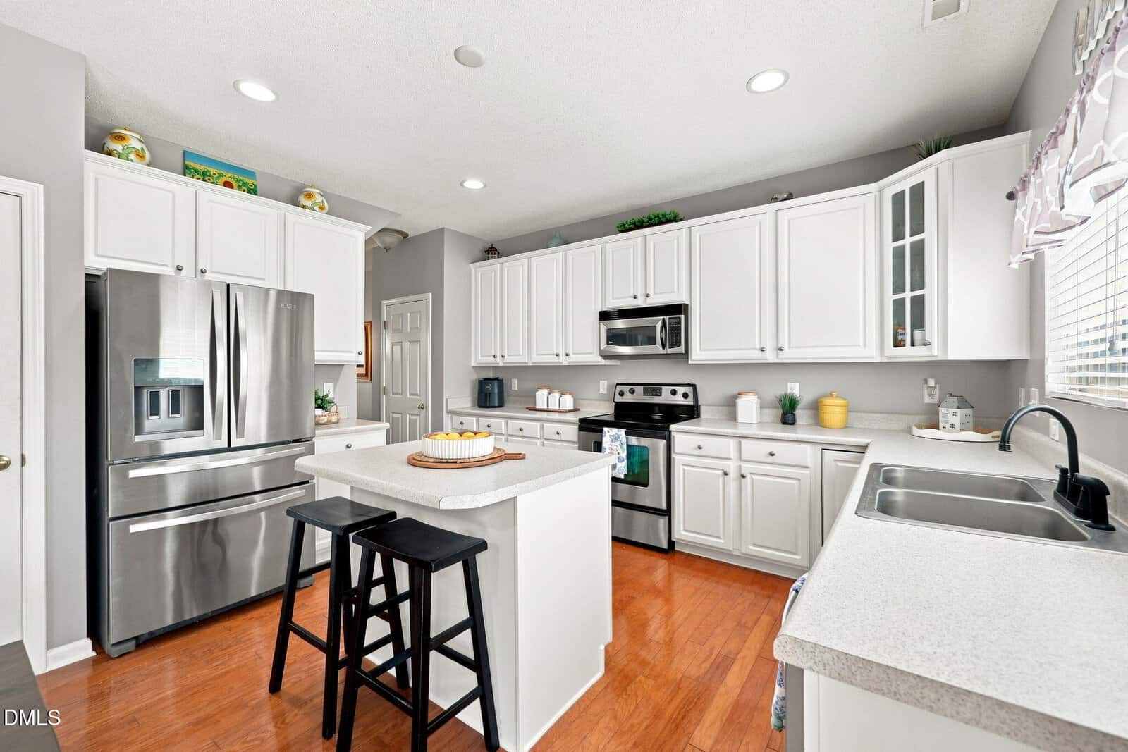 Modern kitchen with white cabinets, stainless steel appliances, small center island with two black stools, wooden floor, and bright natural light from a window with a sheer curtain.