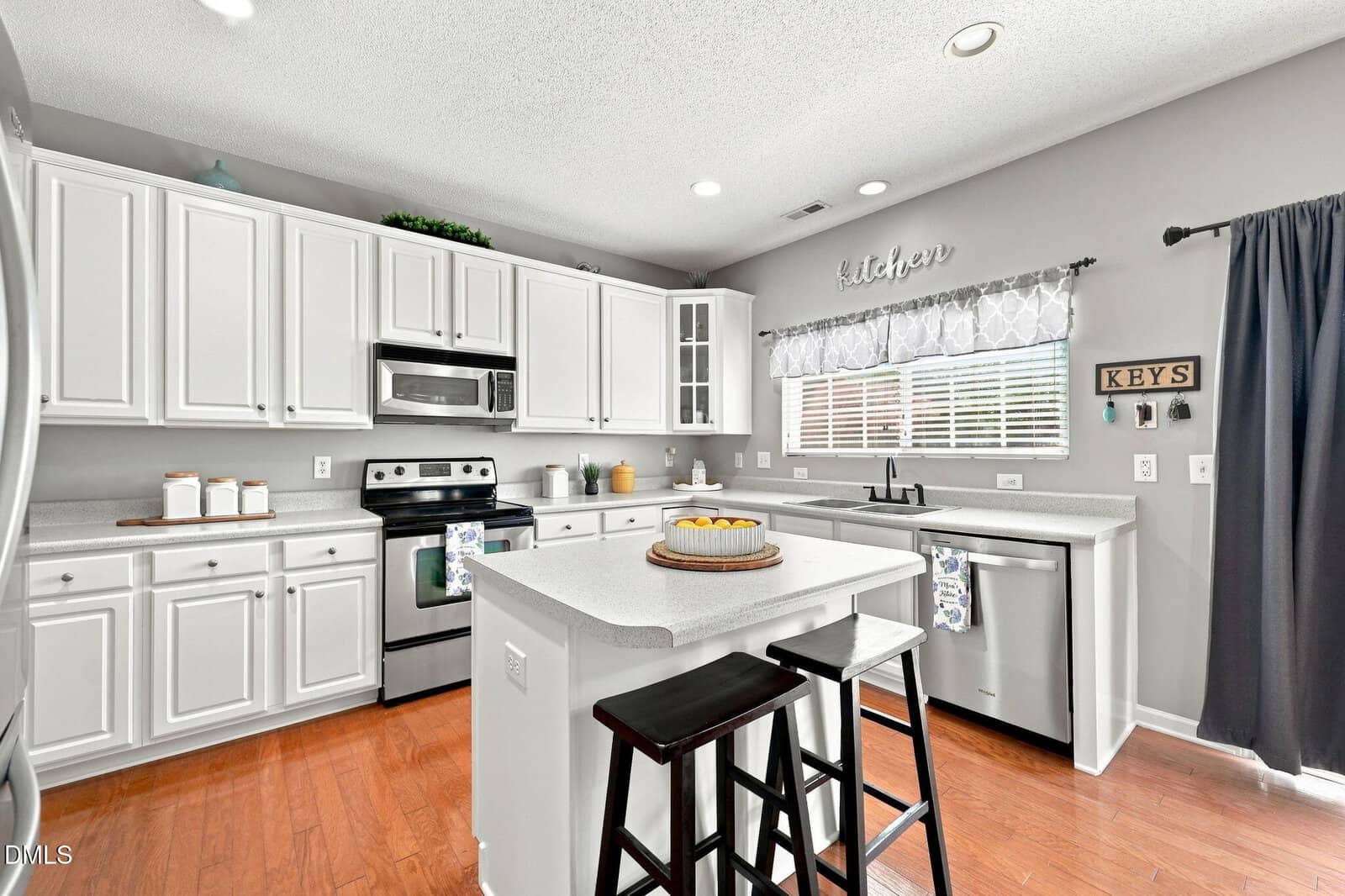 Bright kitchen with white cabinets, stainless steel appliances, a center island with two stools, wooden floors, and a window above the sink with a “kitchen” sign and keys rack on the wall.