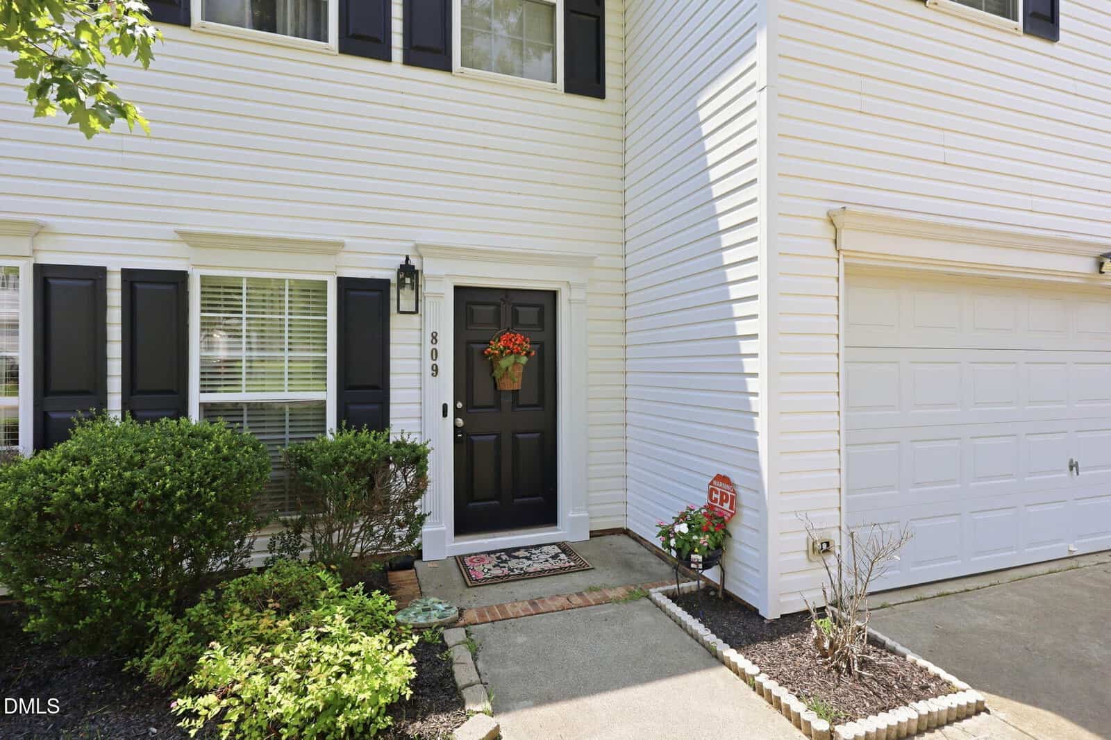Front entryway of a white house with black shutters and a black door, decorated with a flower wreath. Bushes and flowers line the walkway, and a garage door is on the right. A security sign is near the steps.