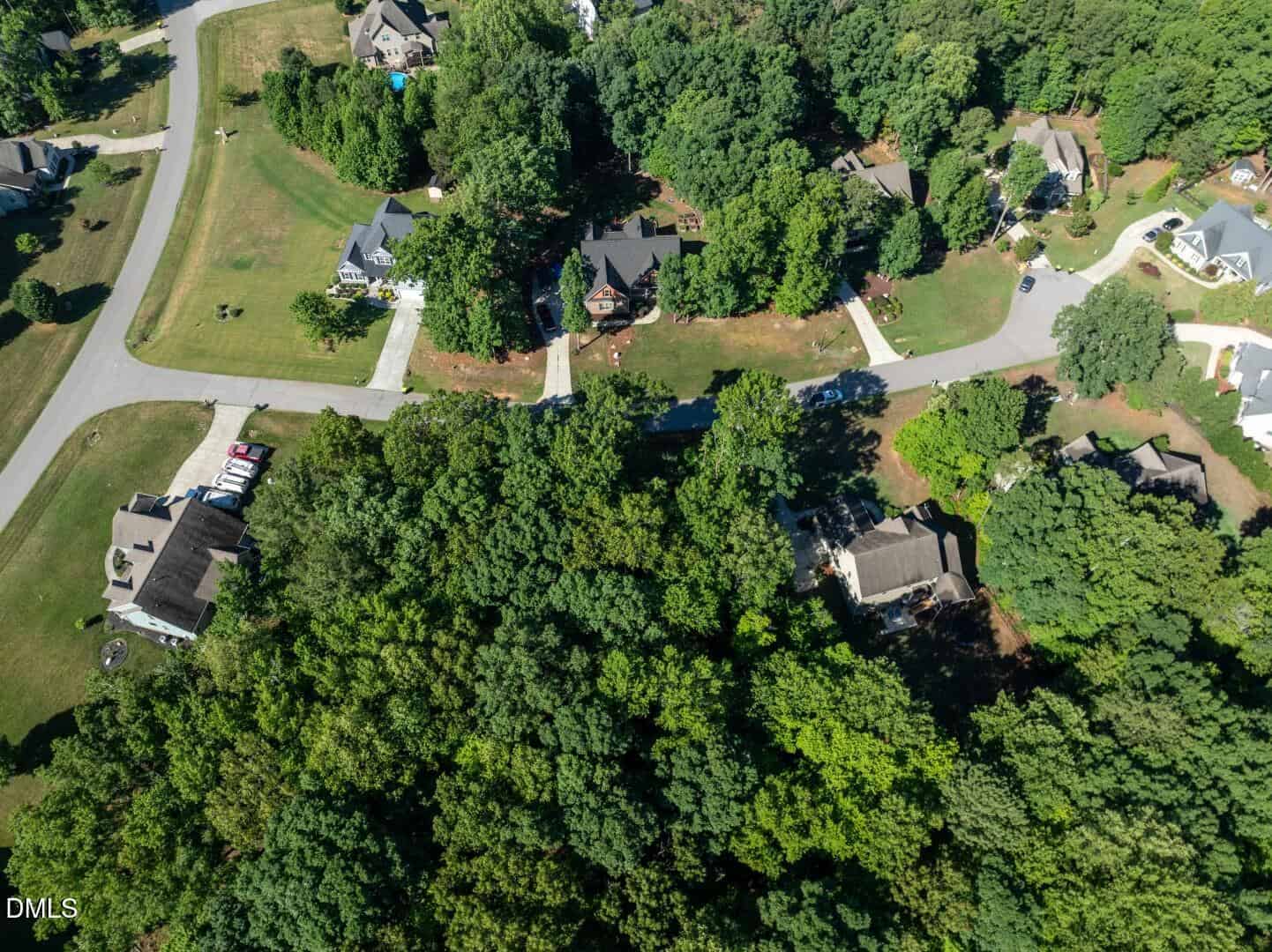 Aerial view of a suburban neighborhood with houses, driveways, lawns, roads, and dense green trees. Several cars are parked near one home. The scene appears calm and residential.