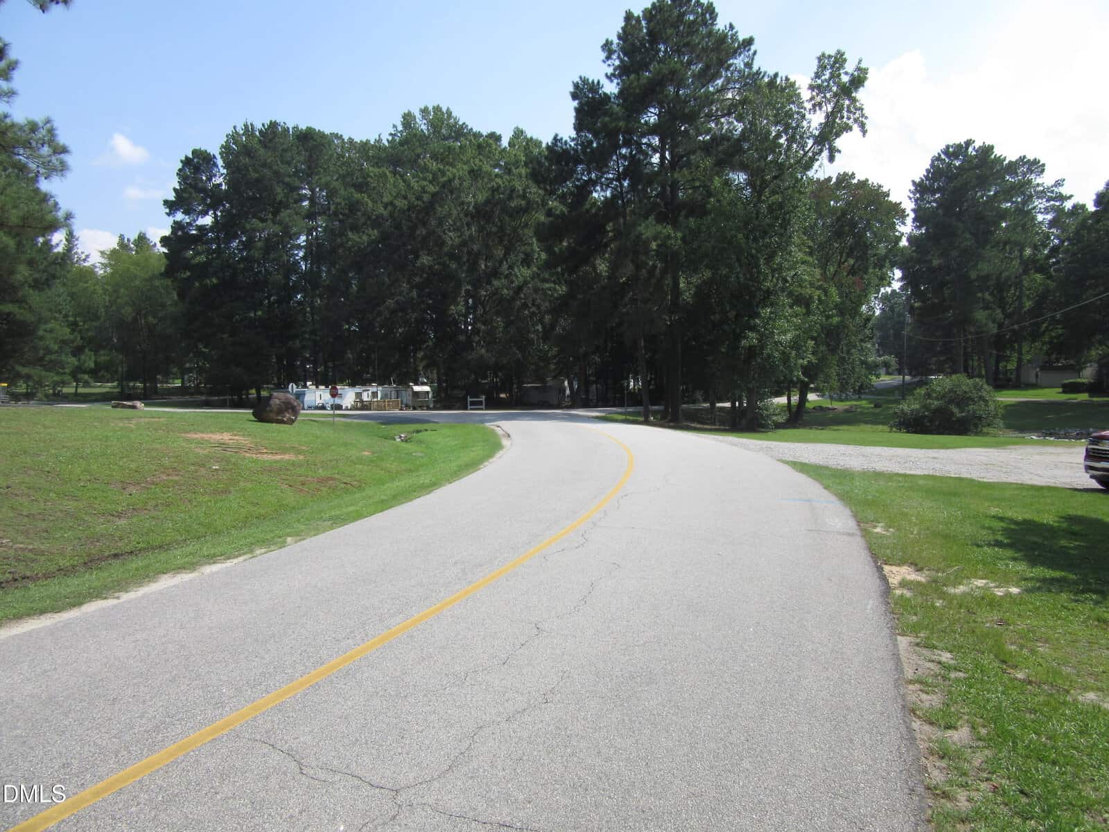 A paved road with a yellow center line curves left through a green, tree-lined area. There are some houses and parked vehicles partially visible in the background among the trees. The sky is mostly clear.