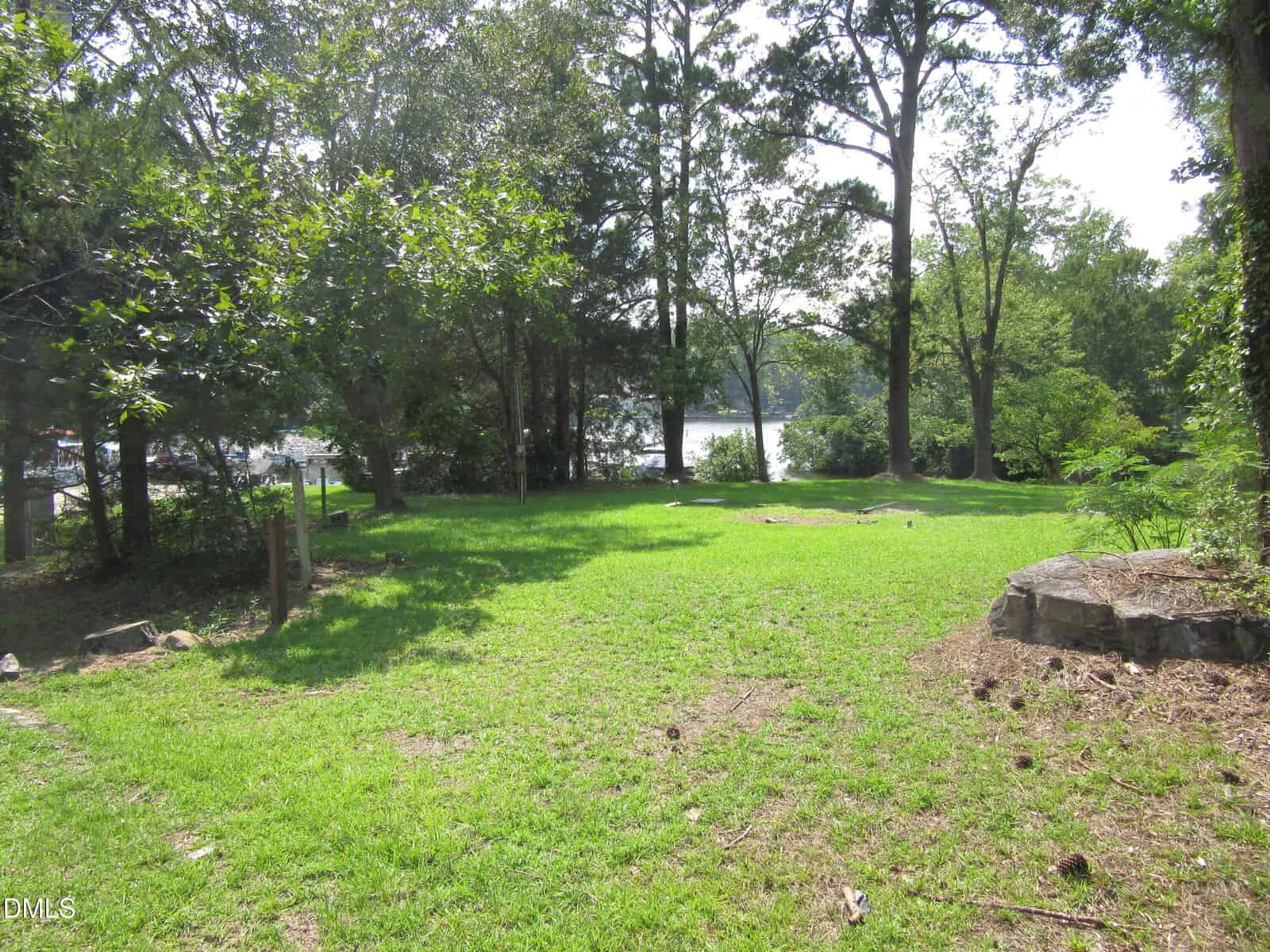 A grassy yard with scattered trees and sunlight filtering through, bordered by rocks on the right side and a view of a lake visible through the trees in the background.