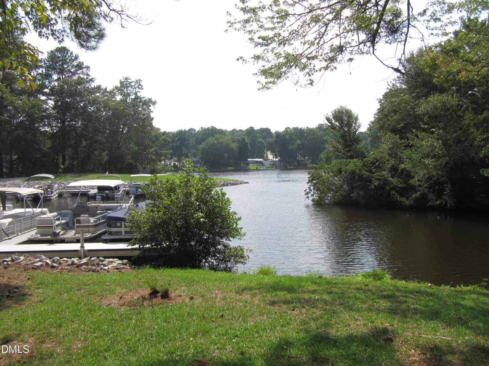 View of a small marina with docked boats on a calm lake, surrounded by green trees and grassy shore under a bright sky.