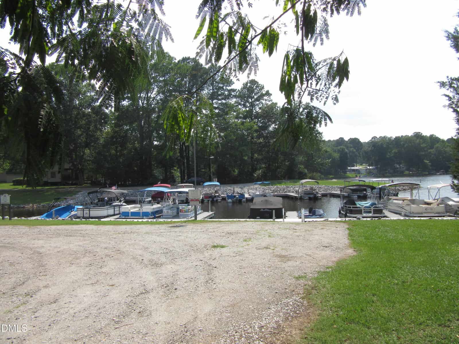 A gravel path leads to a small marina with several boats docked along the water. Trees and greenery surround the area, and some branches hang into the top of the image.