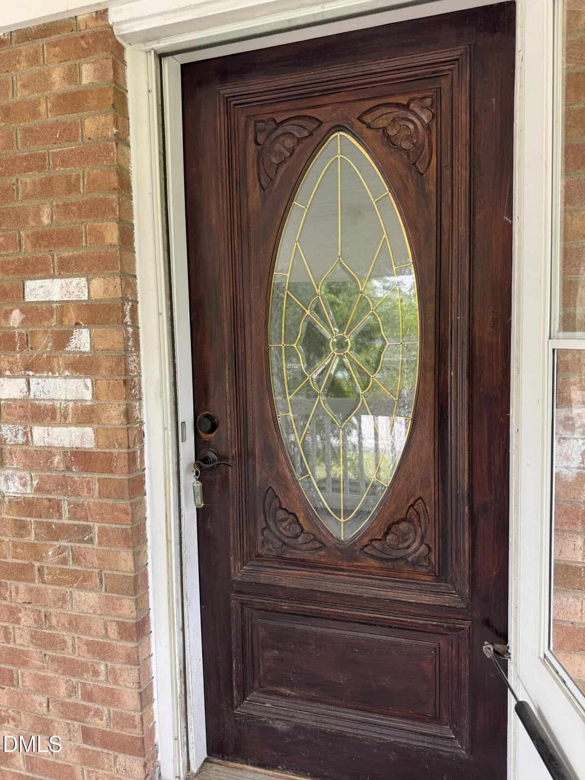 A dark wooden front door with an oval glass panel featuring decorative gold and clear glass designs, set in a brick wall with a white door frame.