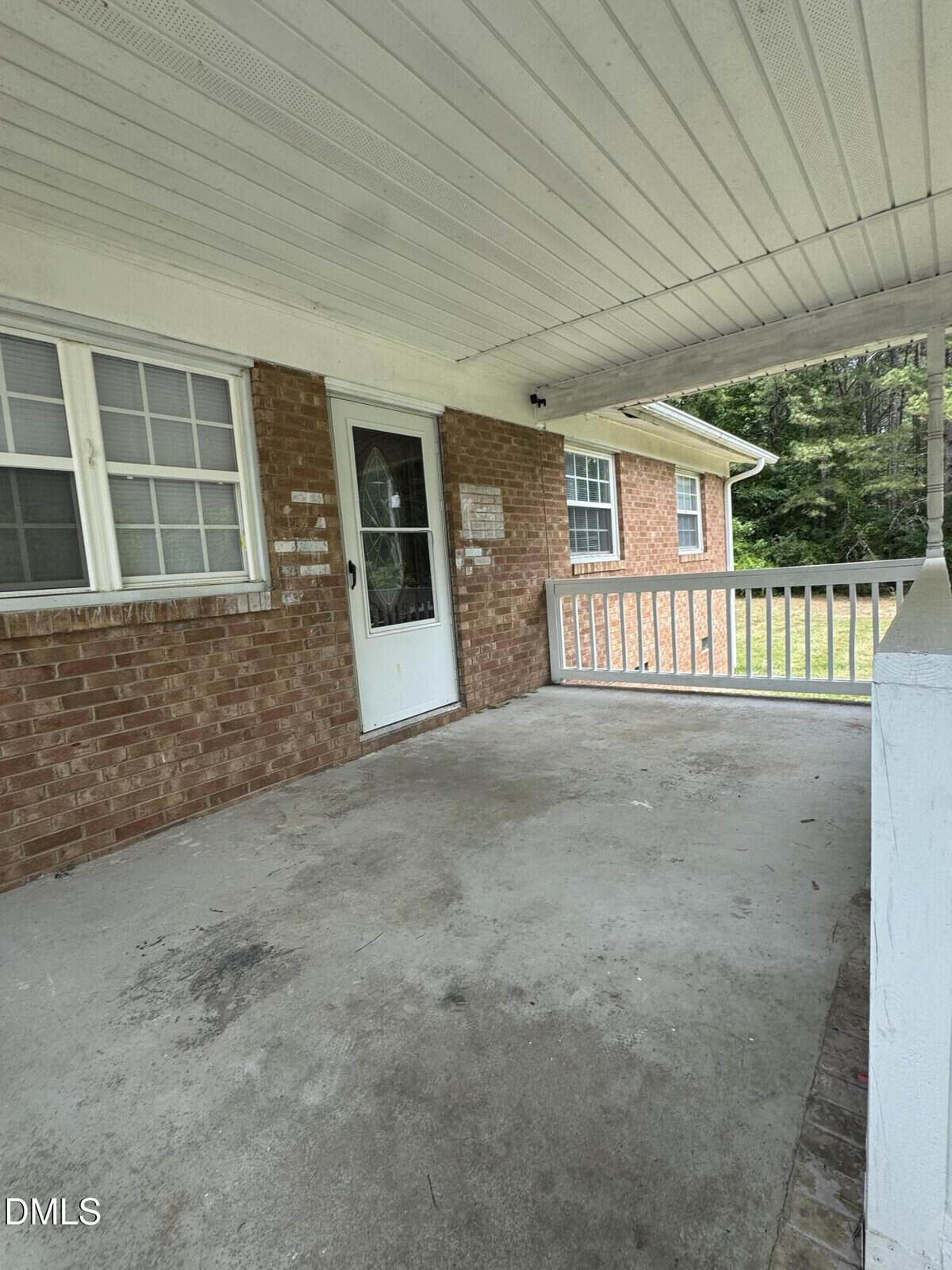 Covered porch with a concrete floor, white railing, brick walls, a white door with a window, and double windows. Green grass and trees are visible in the background. Ceiling features a simple, grooved design.
