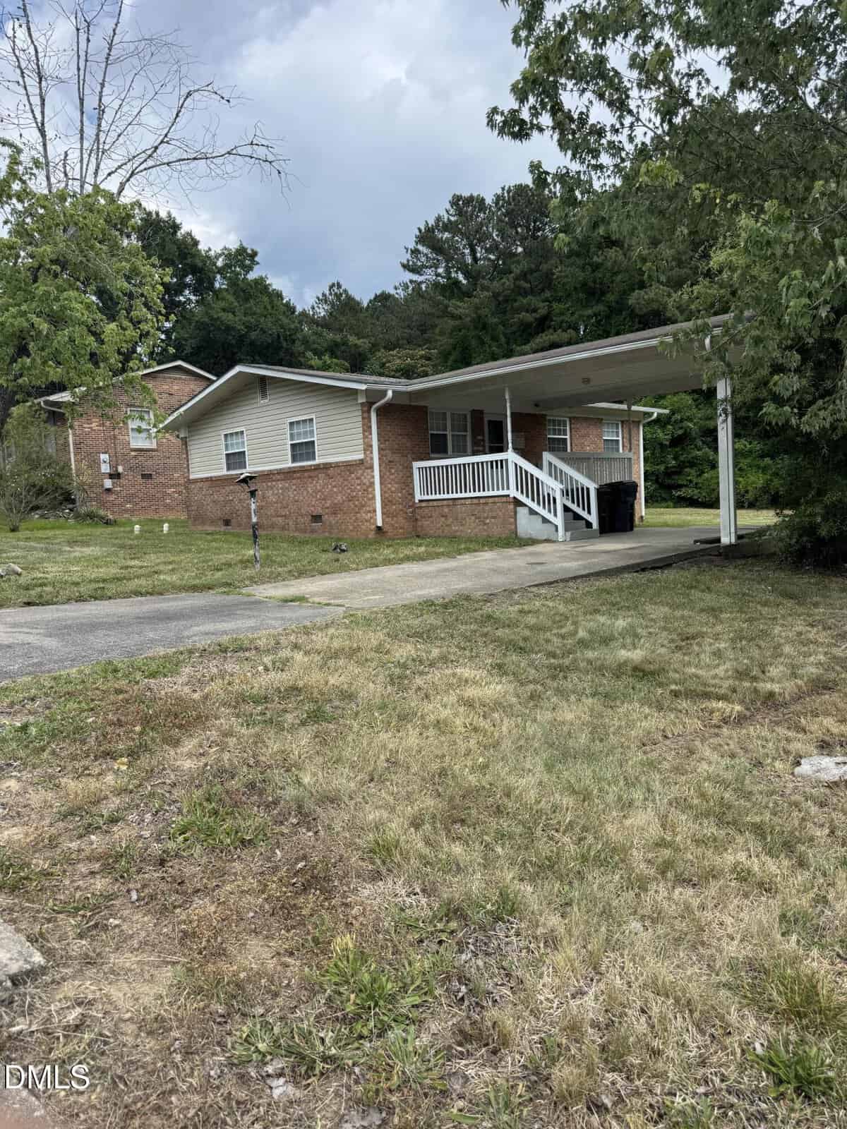 Single-story brick house with beige siding, front porch with white railing, carport on the right, mailbox by driveway, patchy lawn, and trees in the background under a cloudy sky.